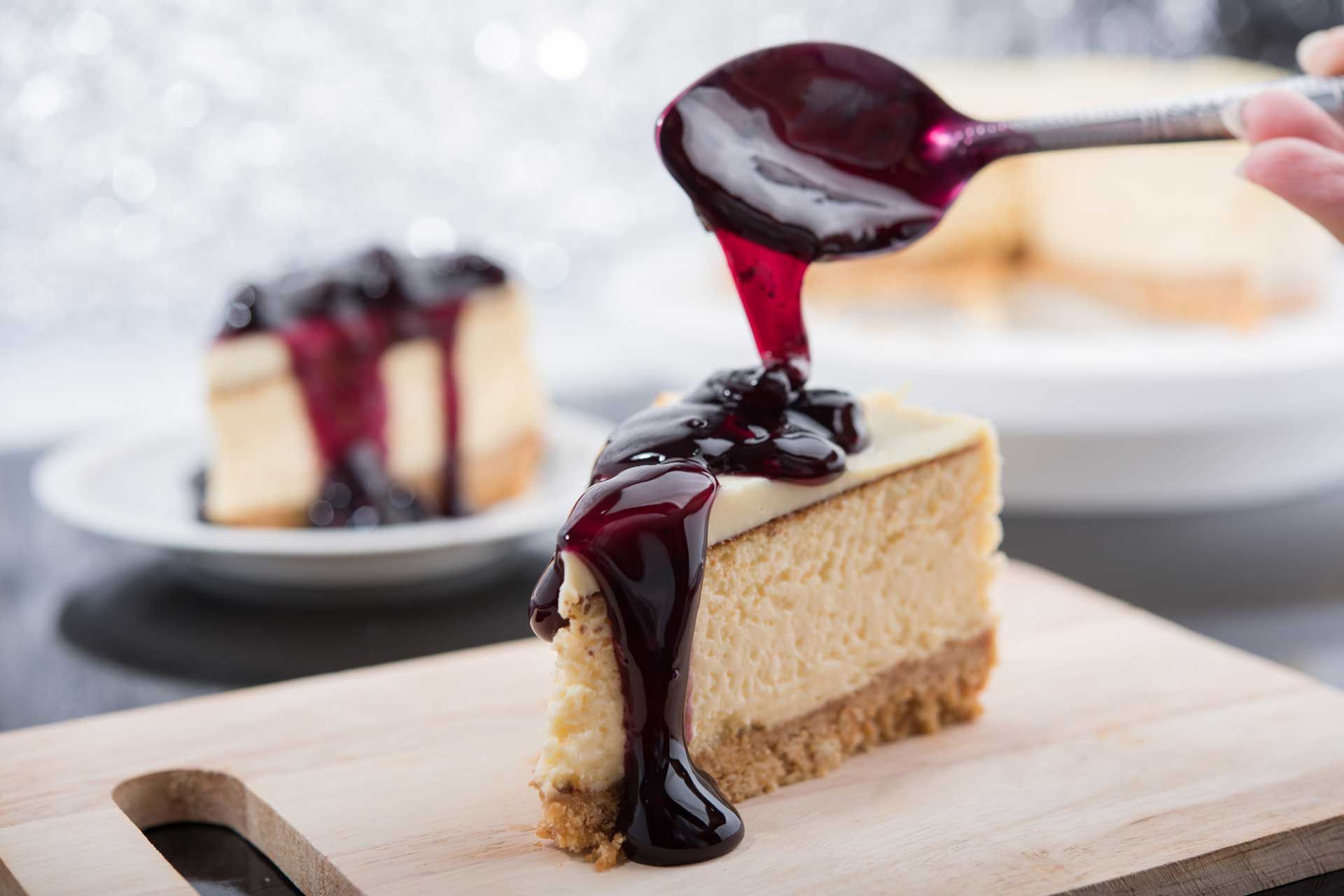 Cheesecake slice with blueberry sauce being poured from a spoon onto it, on a wooden board.