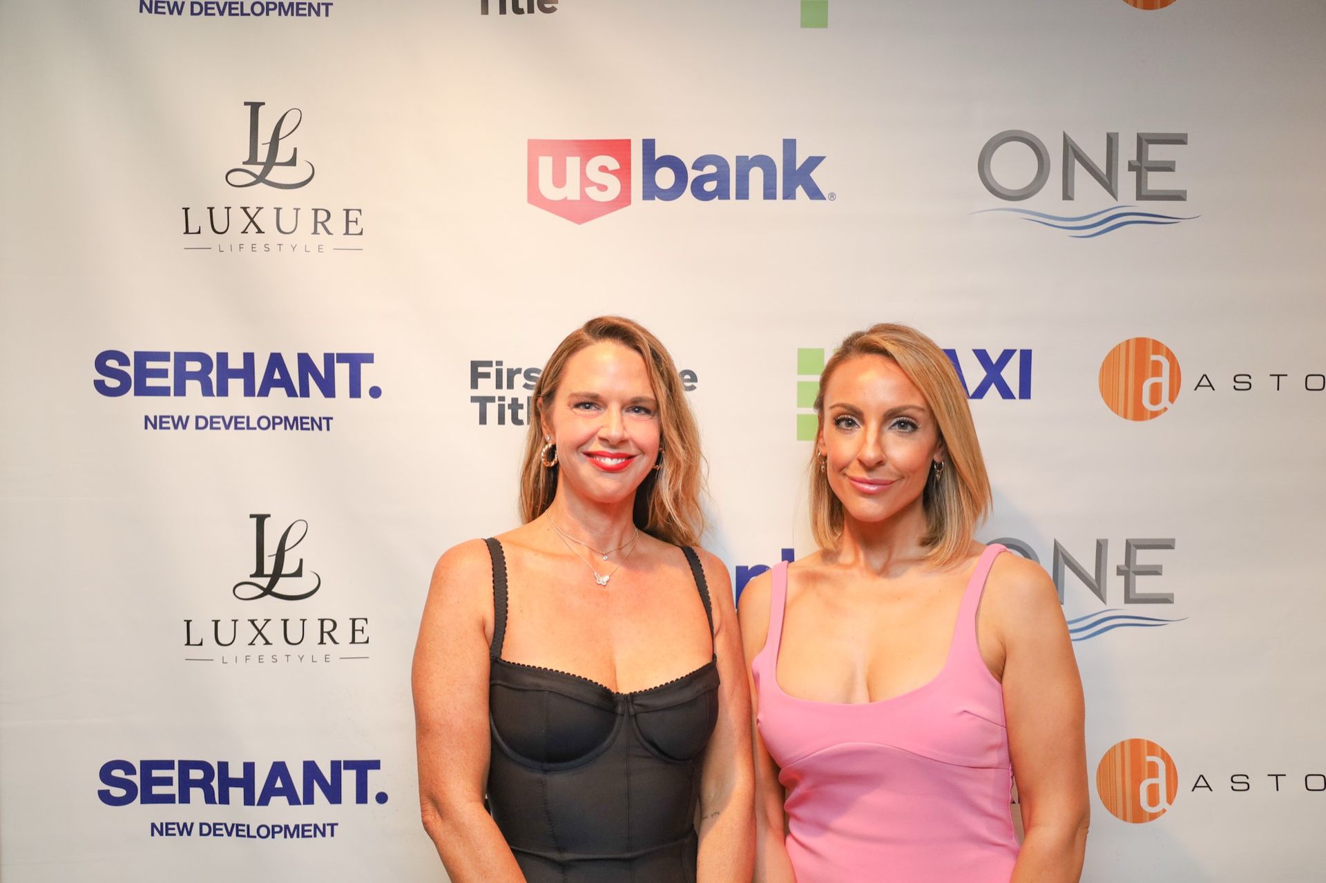 Two women in front of a backdrop with company logos. One wears a black dress, the other a pink top.