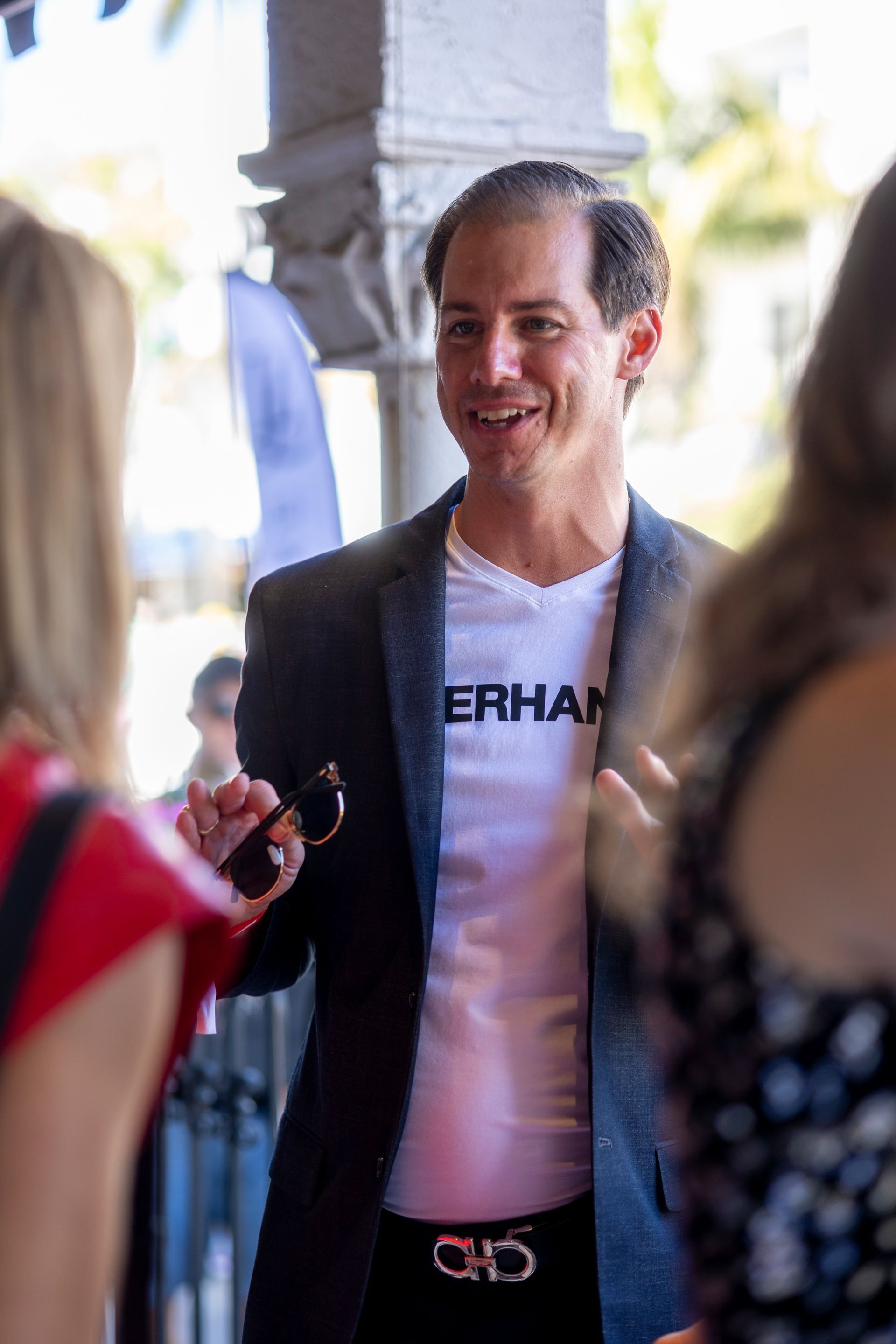 Man in black blazer and white shirt with logo, holding sunglasses, talking to two women.
