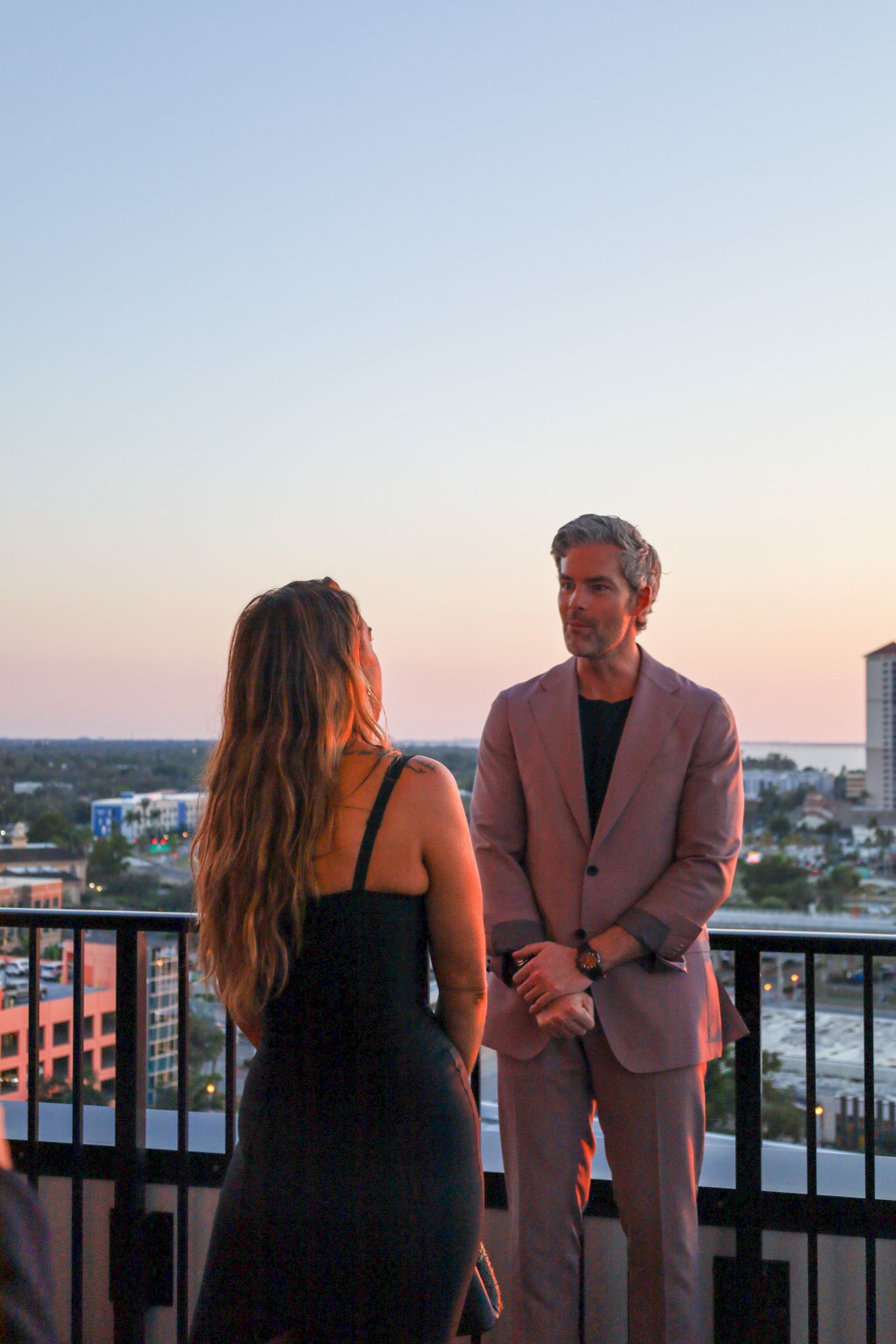 Woman and man stand on a rooftop at sunset. Man in a pink suit, woman in a black dress, both facing each other.