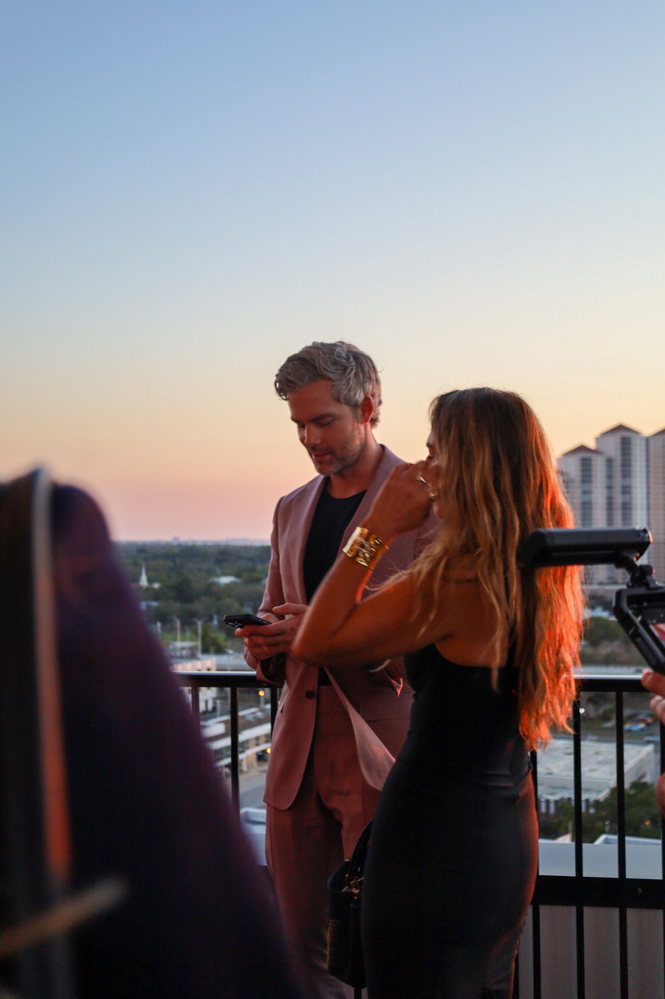 Two people on a rooftop at sunset. Man in a suit looks at his phone, woman in a black dress looks off.