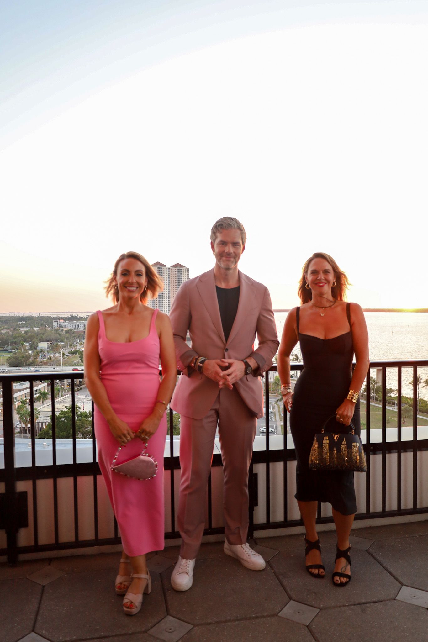 Three people posing outside with water in the background; a person in a pink suit, a woman in a pink dress, and a woman in a black dress.
