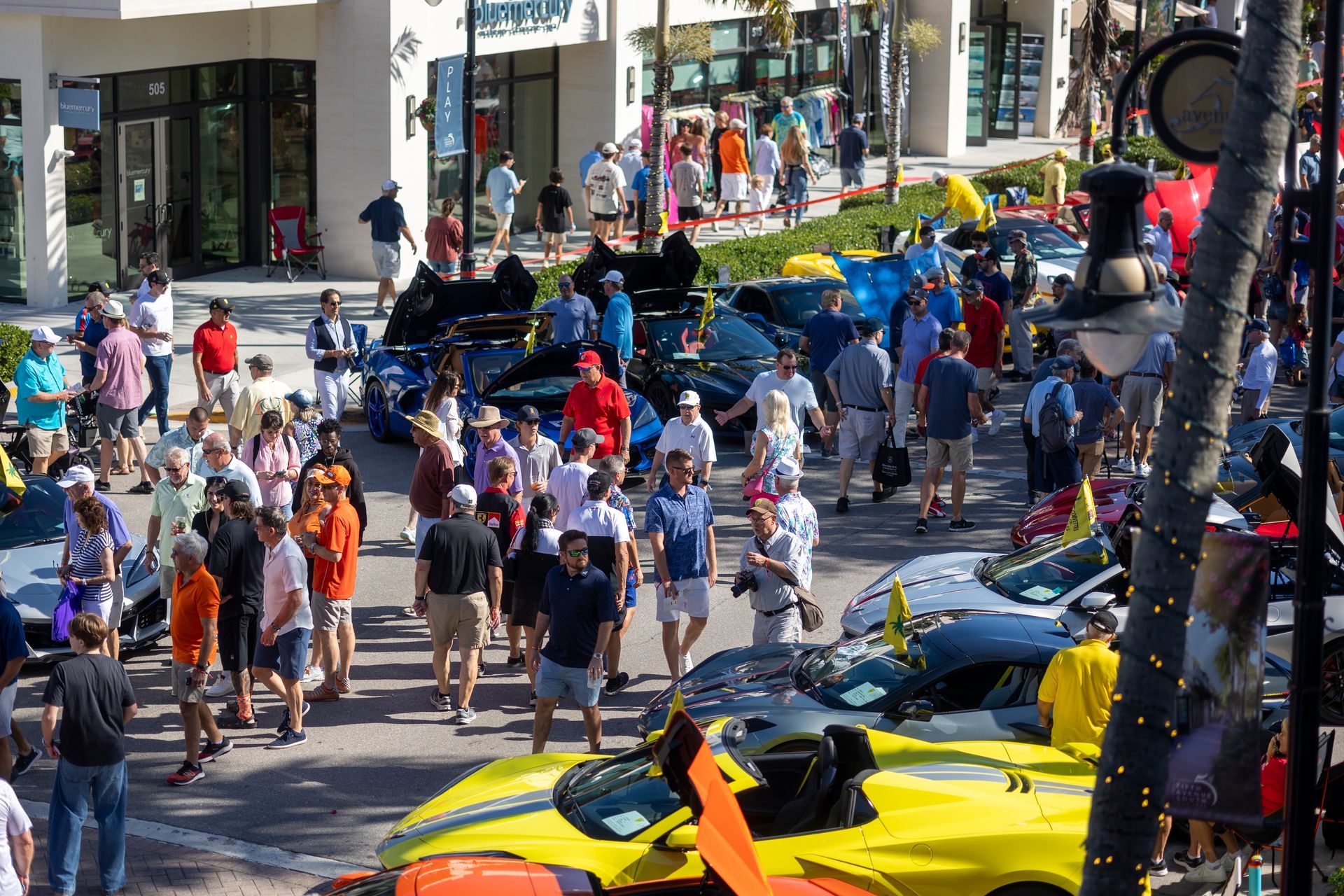 Crowd viewing sports cars on a sunny street. People in casual clothes walk around the vehicles, some brightly colored.