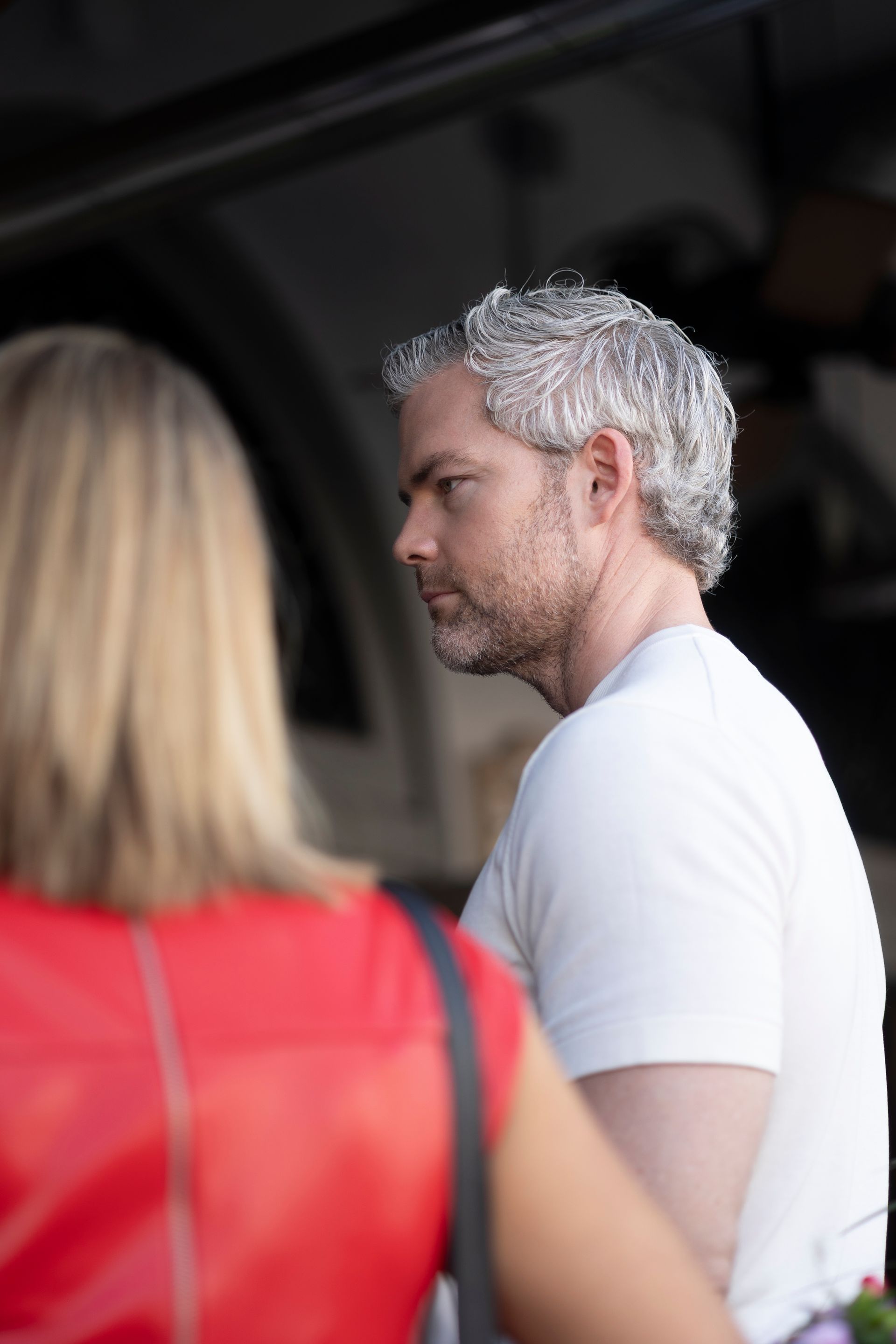 Man with gray hair and beard, wearing a white shirt, looks to the side while standing near a person in a red top.