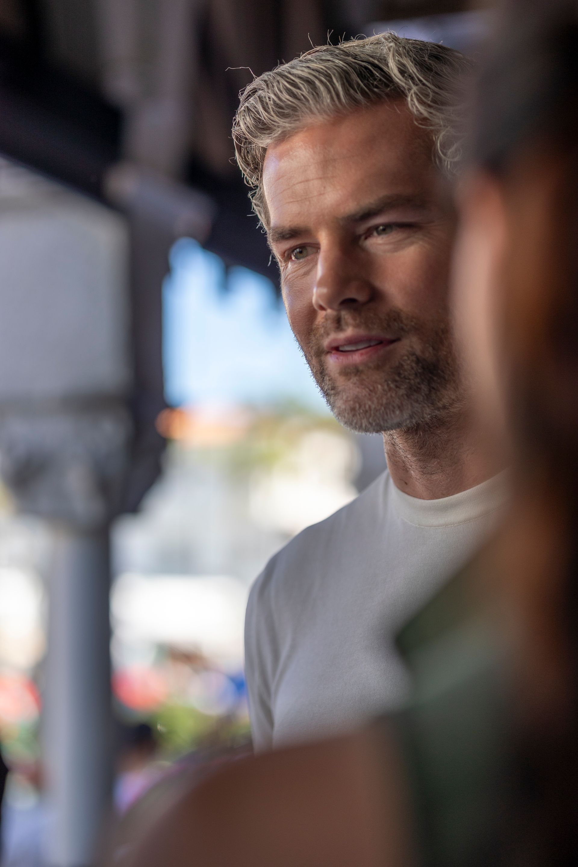 Man with graying hair in white shirt, looking at someone off-screen in a bright outdoor setting.