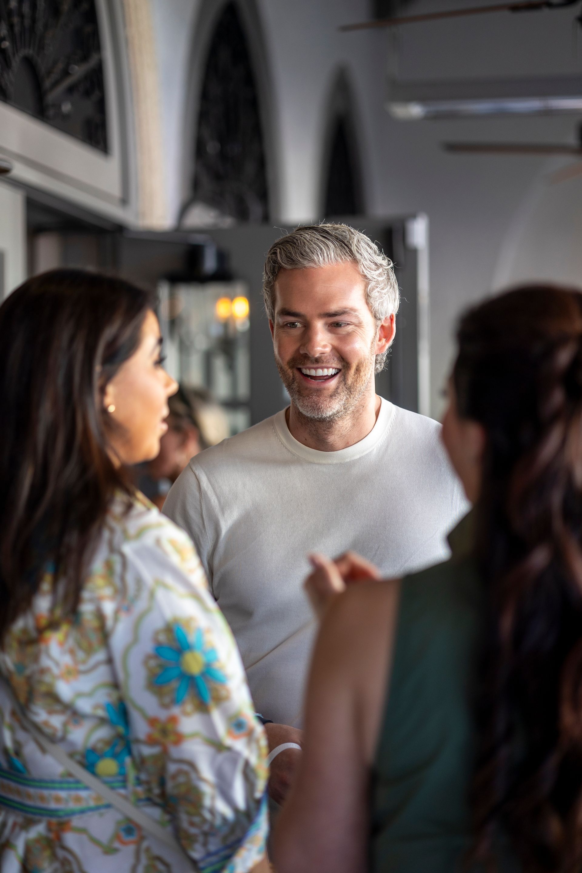 Man smiles while talking to two women in a bright indoor setting.