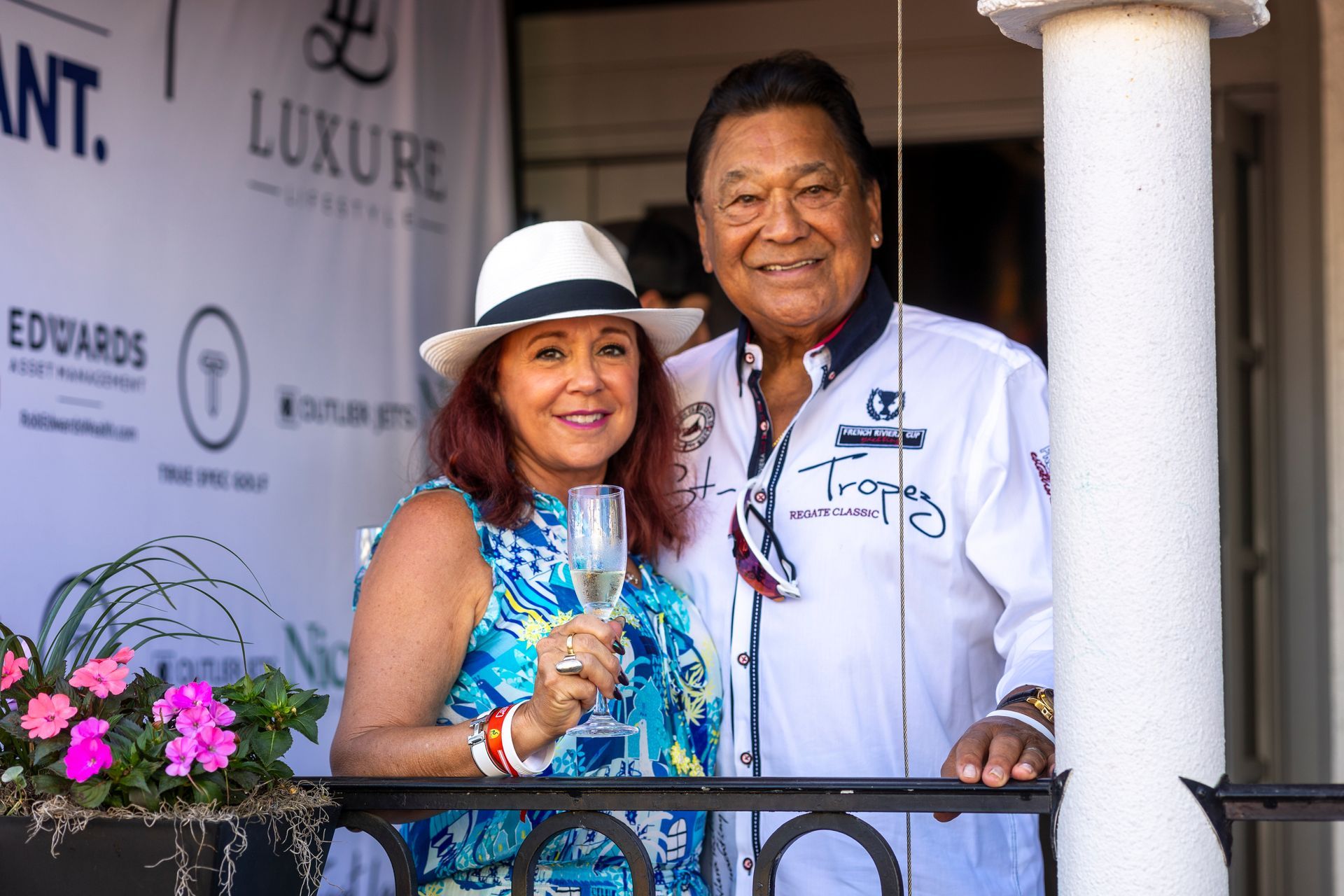 Couple on a balcony, woman in blue dress holding champagne, man in white shirt, brands in the background.