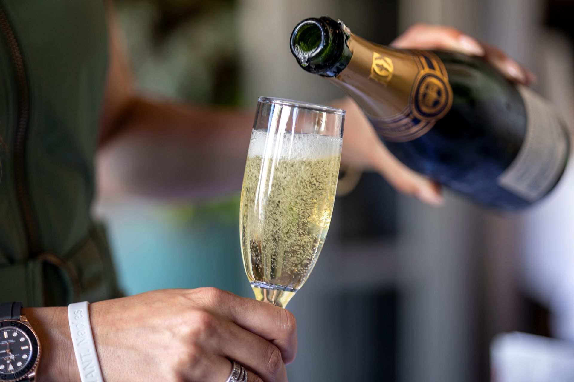 Pouring champagne into a flute; bubbles visible. Person in green holding glass and bottle with watch on wrist.