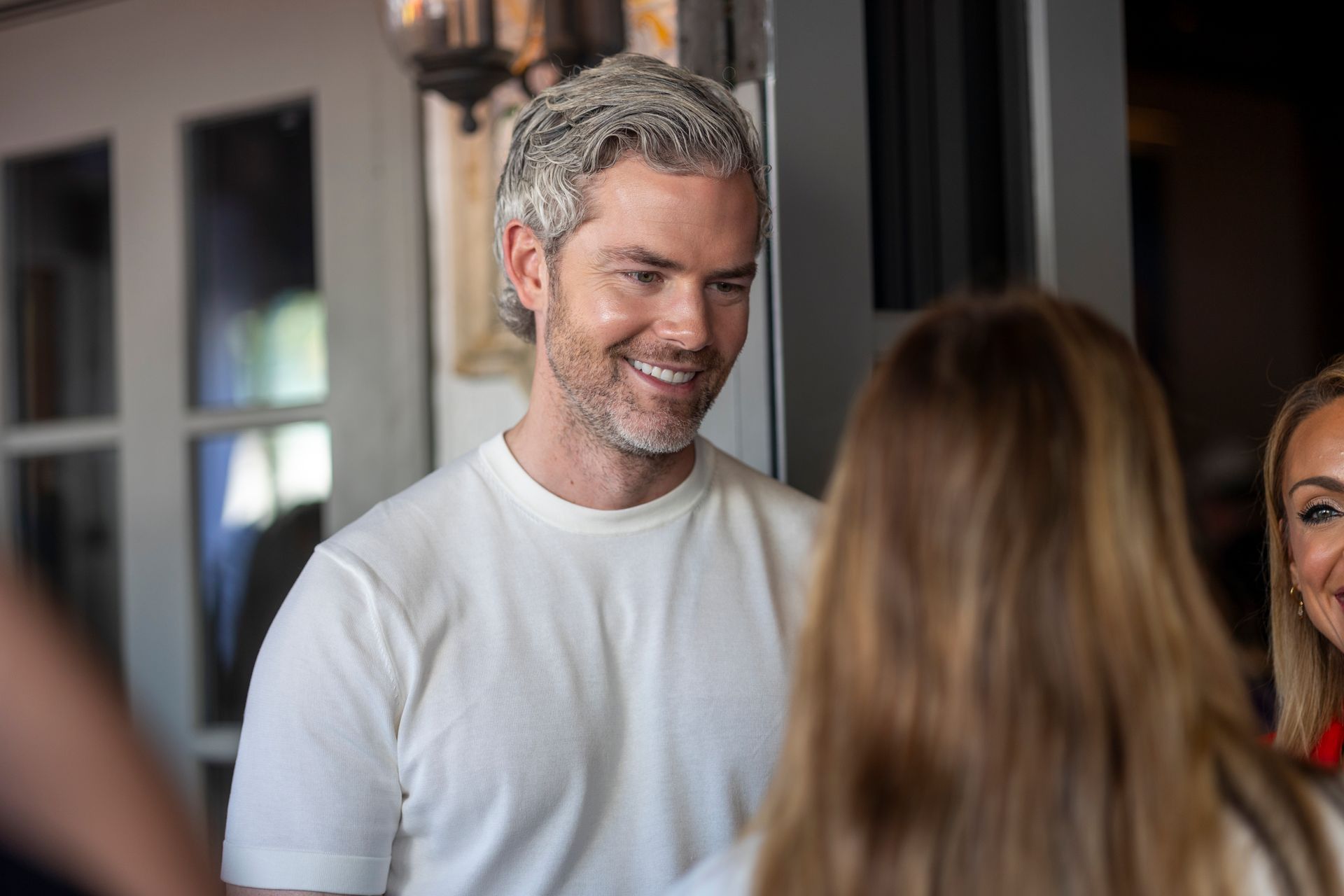 Man with graying hair smiles, talking to two people indoors.