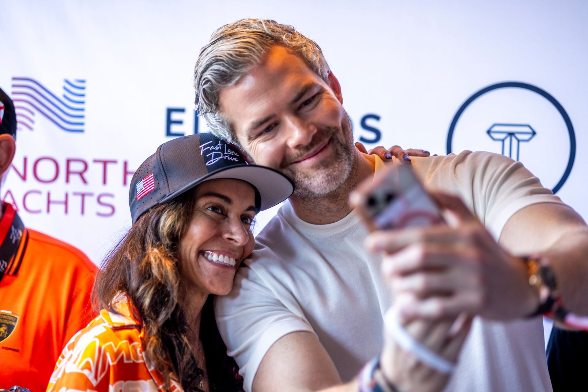 Smiling man and woman taking a selfie; woman in a hat, man has gray hair; white background, logos.