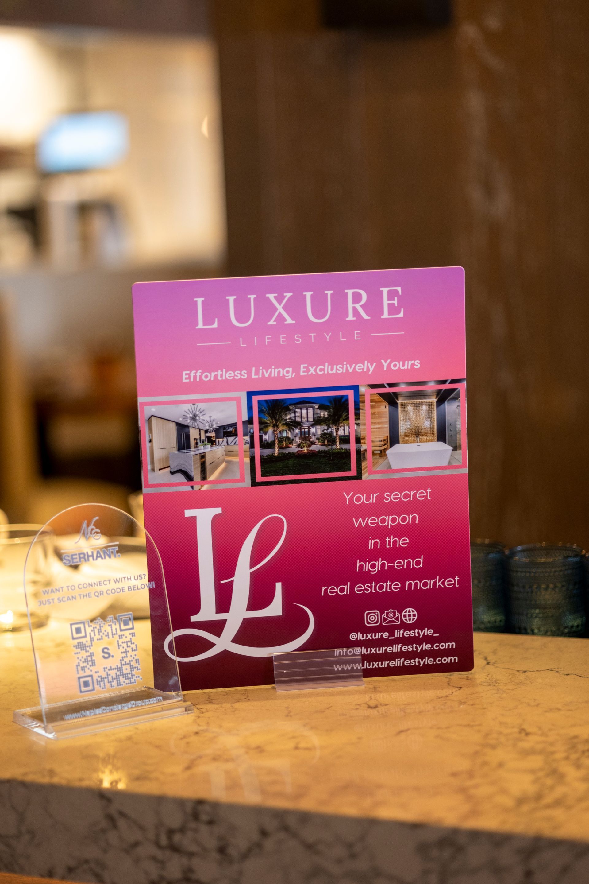 A pink and purple Luxure Design sign on a counter with photos of rooms and the letter