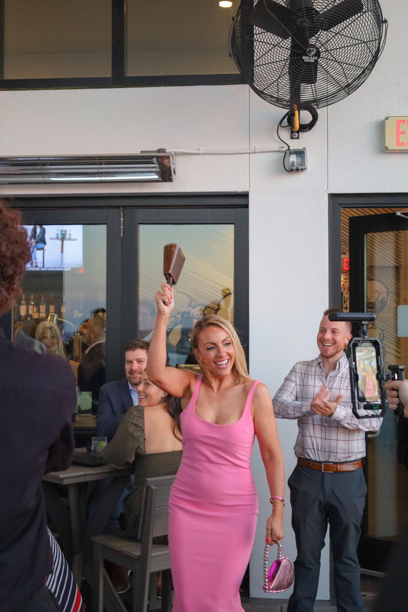 Woman in pink dress celebrates, holding a purse and wooden object, outside a restaurant. A man claps nearby.