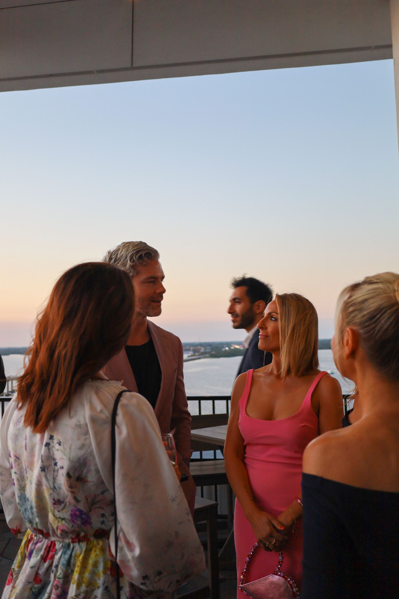 Group of people socializing on a balcony at dusk, with water visible in the background.