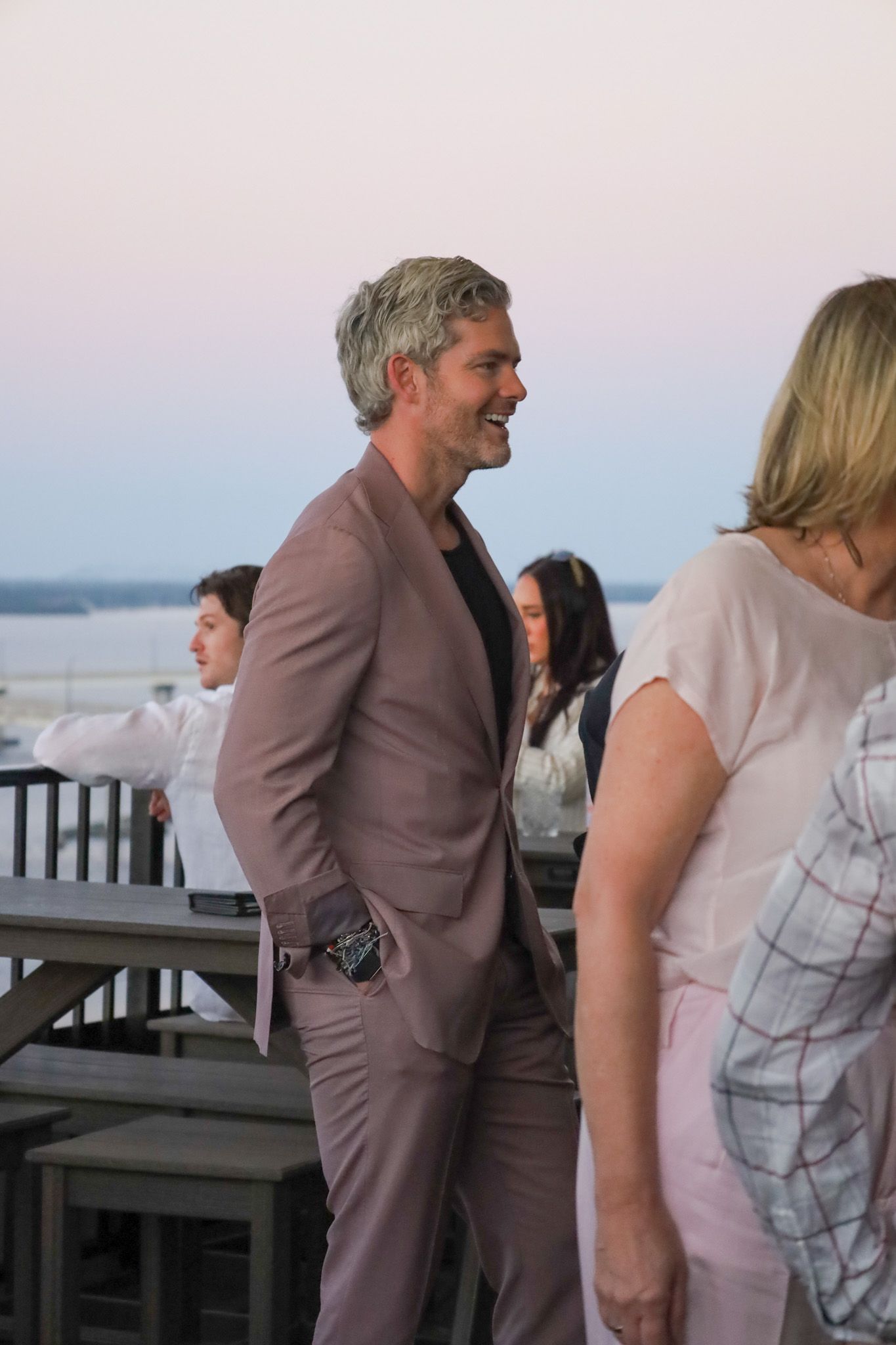 Man in a mauve suit smiles outdoors, hand in pocket.  People and a railing are visible nearby.