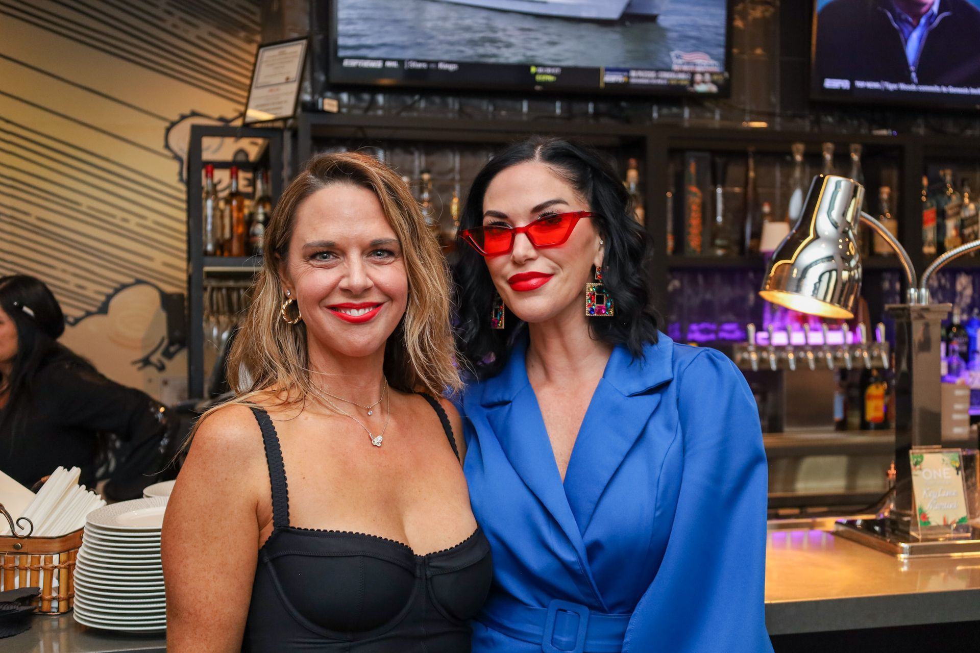 Two women smiling at a bar. One wears a blue blazer and red sunglasses; the other, a black top.