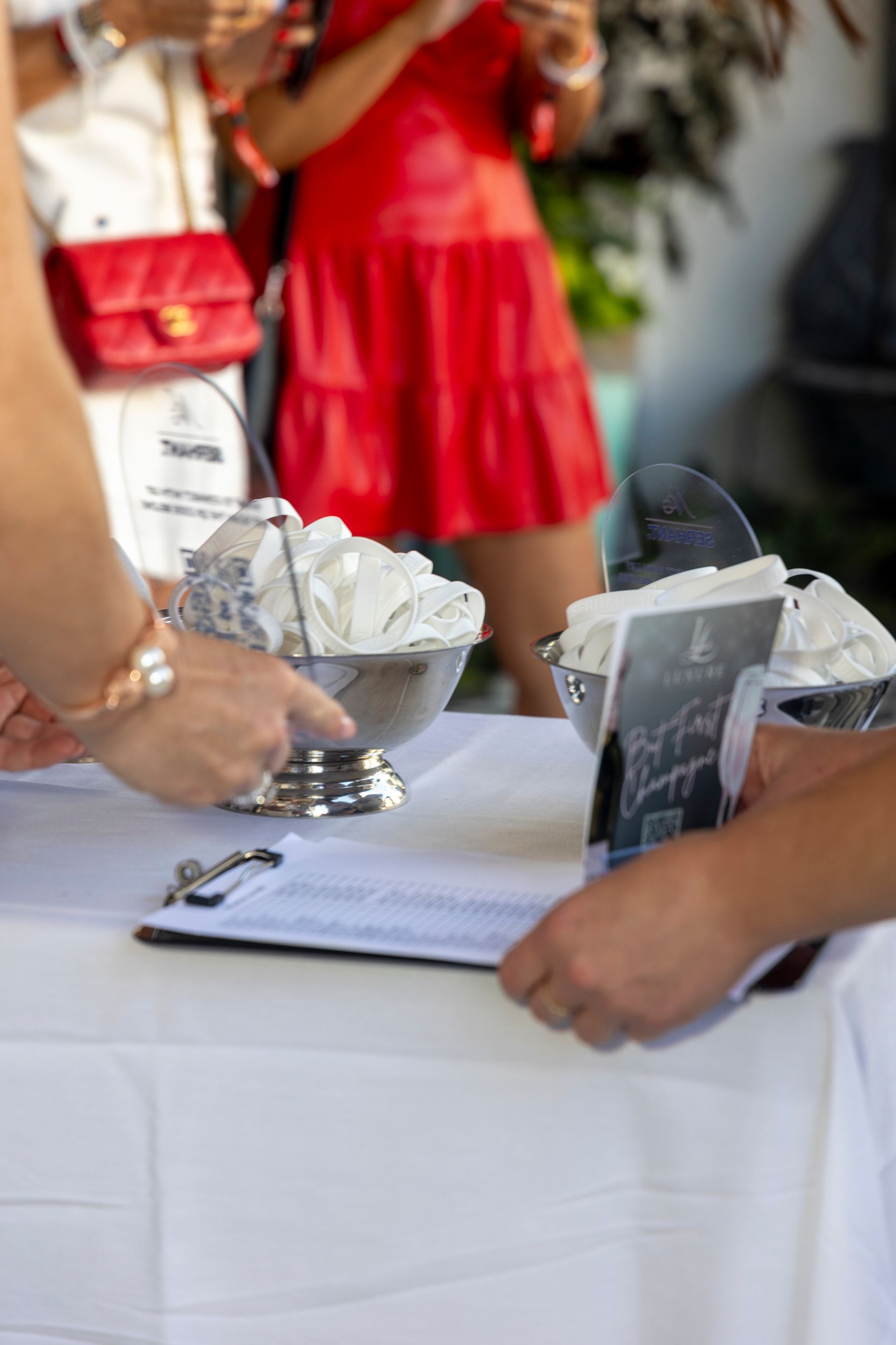 People at a table, silver bowls with napkins, clipboard, and paperwork. A woman in red dress in background.