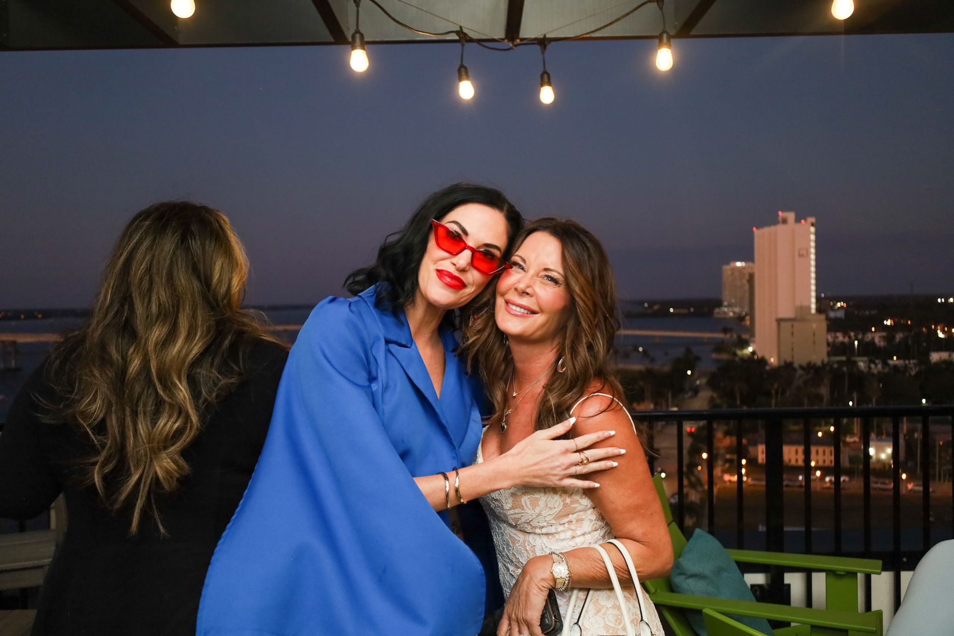 Two women embrace on a rooftop at dusk; one wears red sunglasses and a blue jacket.