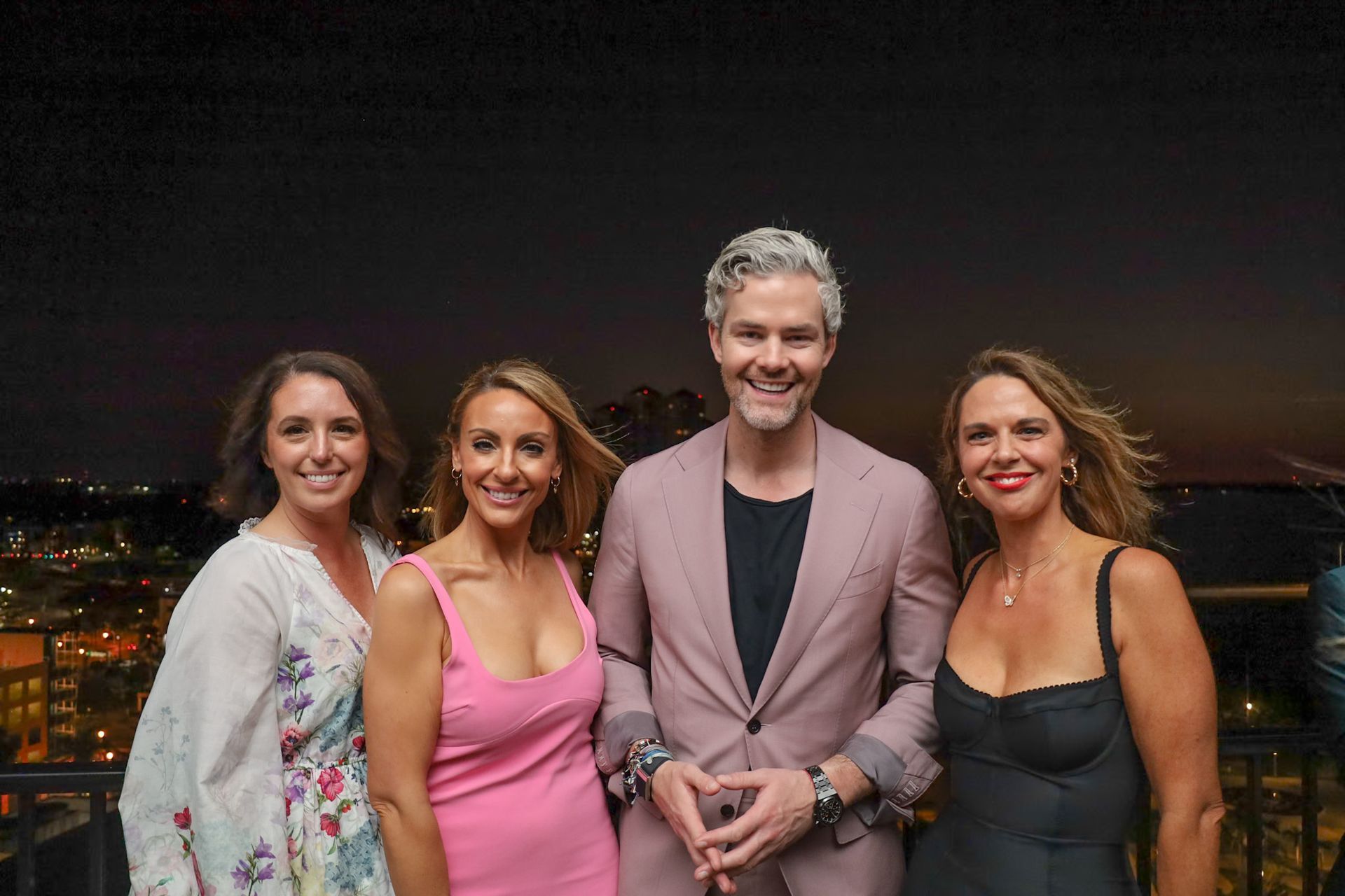 Four people pose outdoors at dusk: a man in a pink suit, and three women in dresses, smiling. City lights in background.