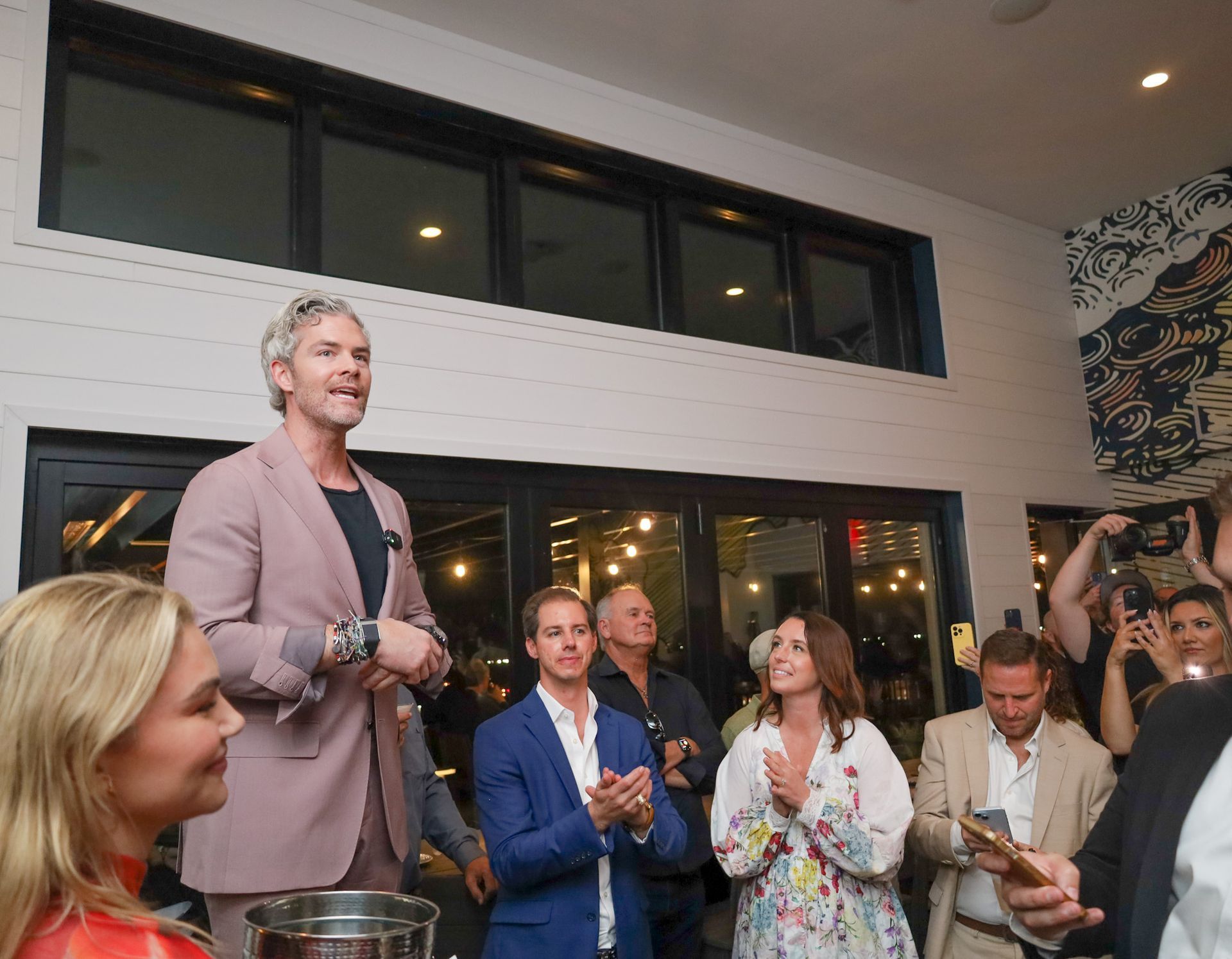 Man in pink suit speaks to a crowd indoors. People clap, some take photos.
