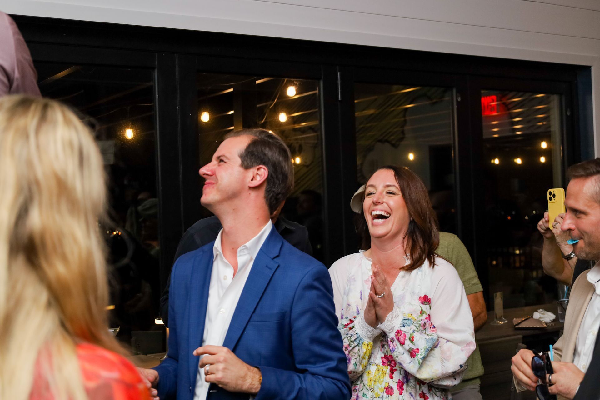 People celebrating. Woman laughing, clapping. Man in blue suit looking up. Lit background.