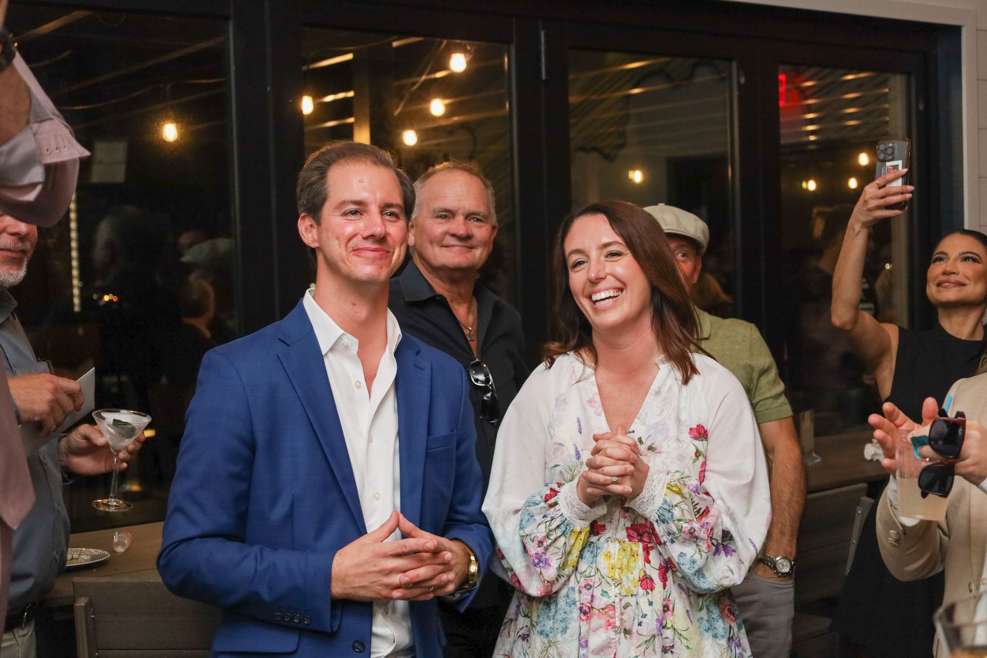 People smiling and clapping indoors. Man in blue blazer, woman in floral top.