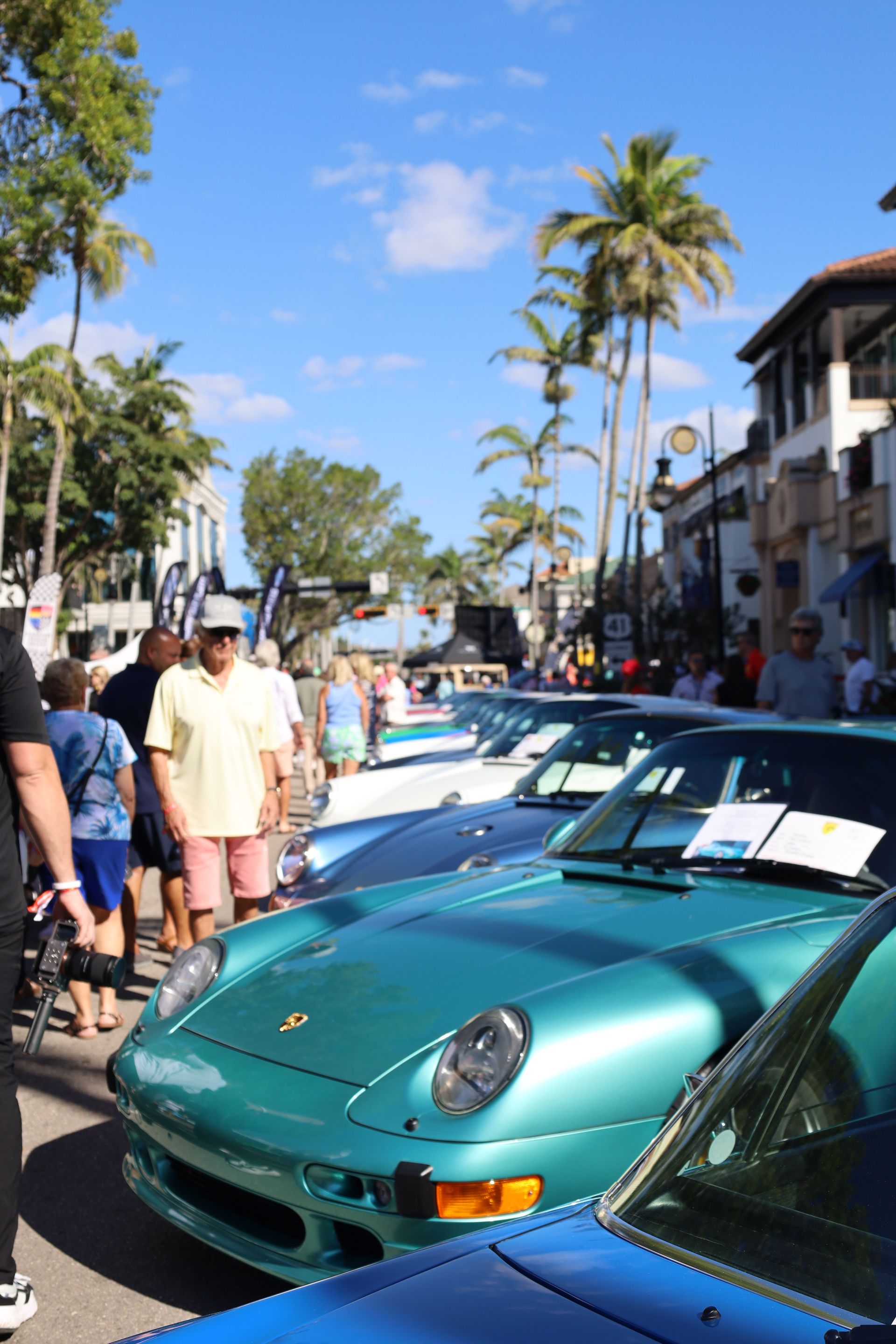 Classic turquoise Porsche and other cars on display at an outdoor car show, palm trees in background.