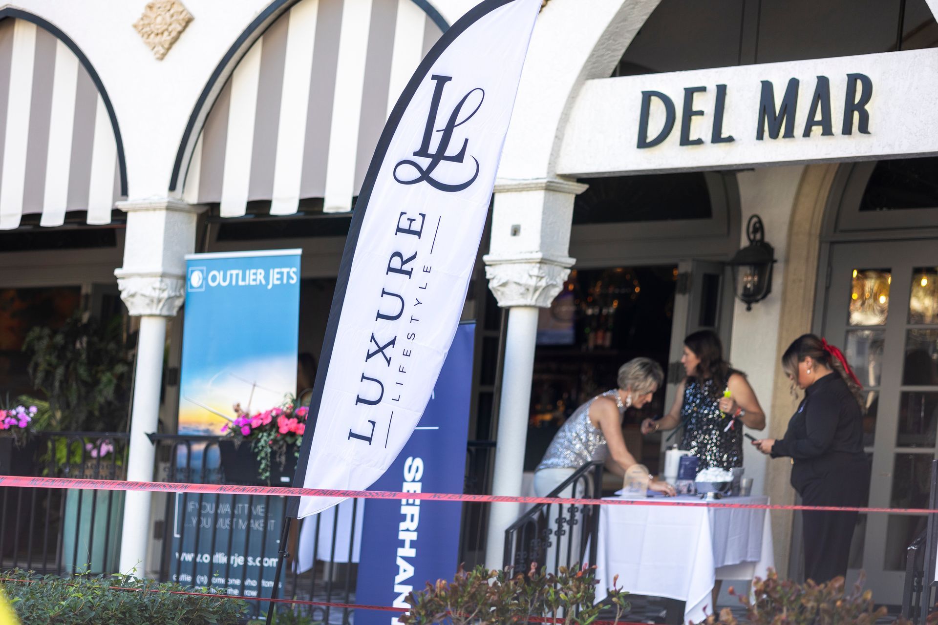 Entrance to Del Mar restaurant with people near a table, signs for Luxure Jets and Serhant.