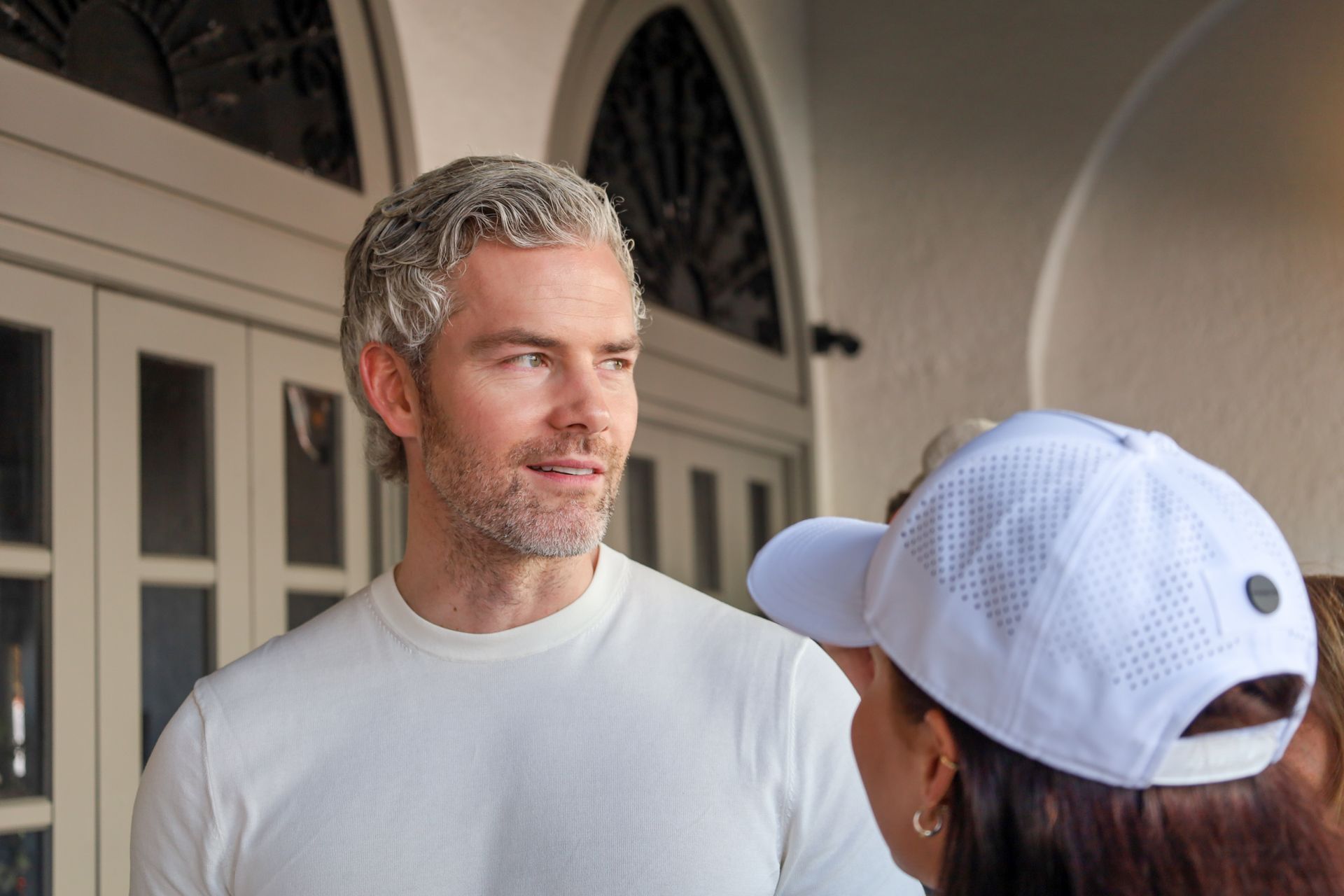 Man with gray hair, white shirt, conversing with person in white hat near arched doorway.