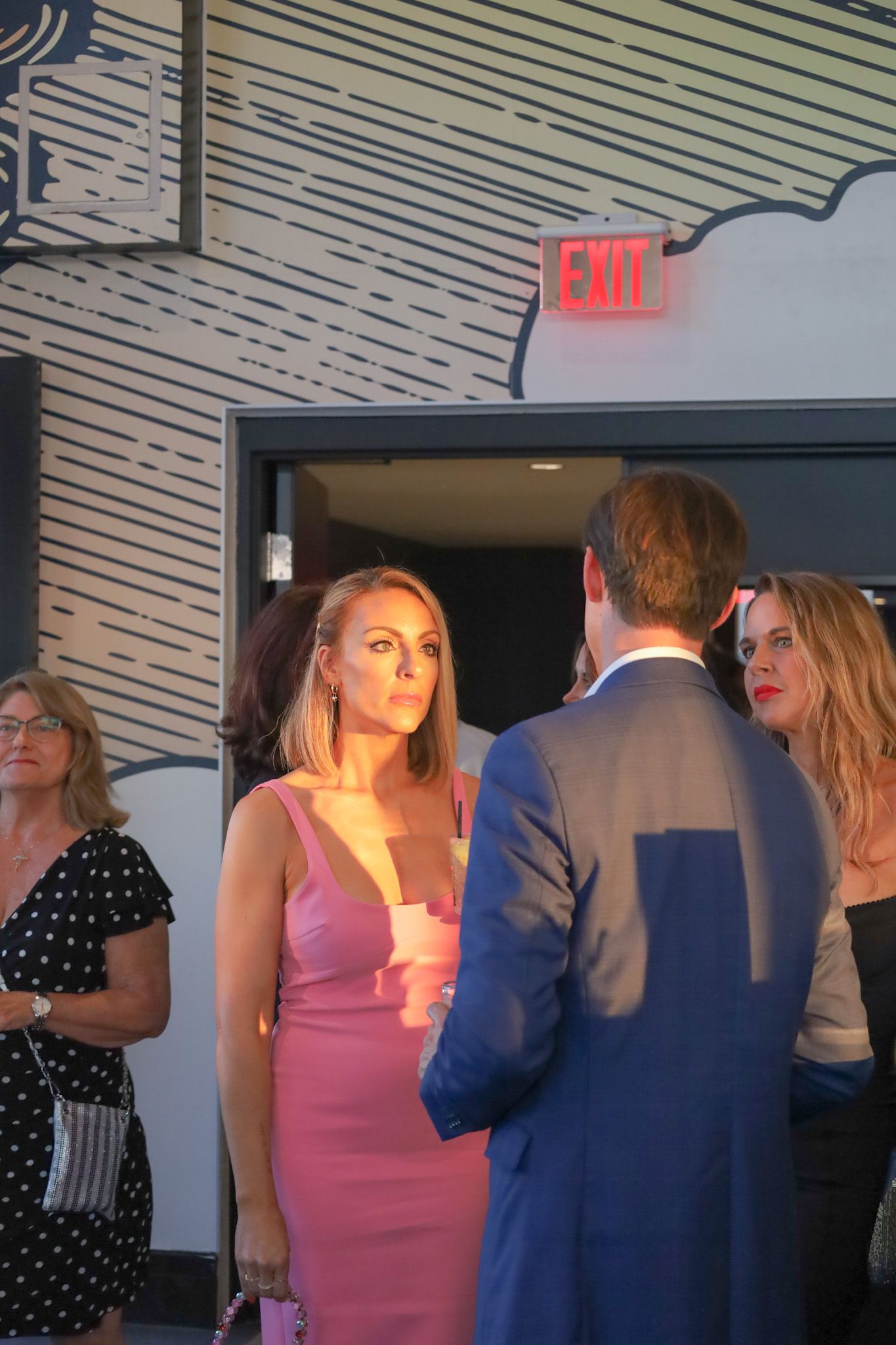 Woman in pink dress talks to man in blue suit, others in background. Interior setting.