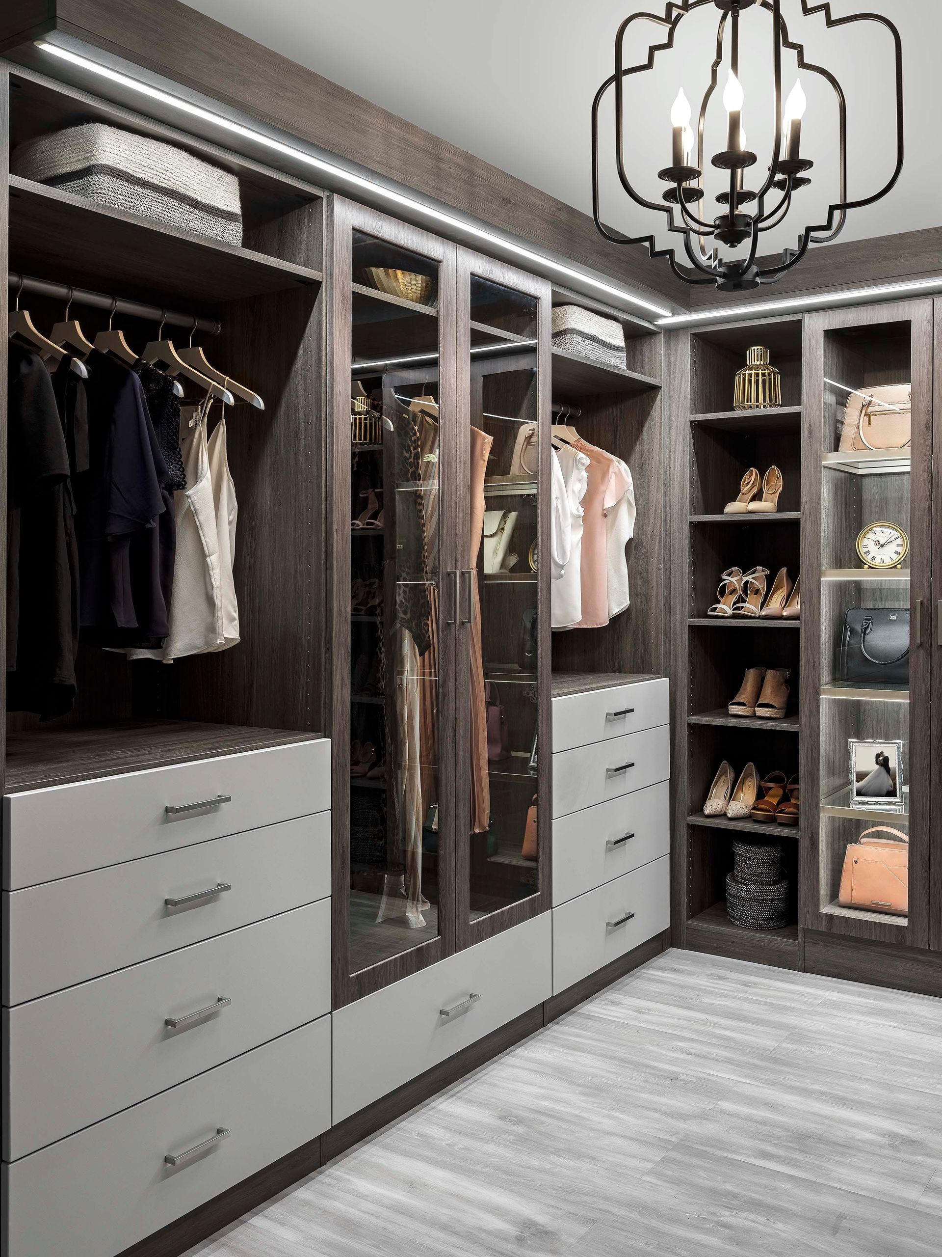A walk-in closet featuring dark wood cabinetry, grey drawers, glass-front cabinets, open shelving, and a metal chandelier.