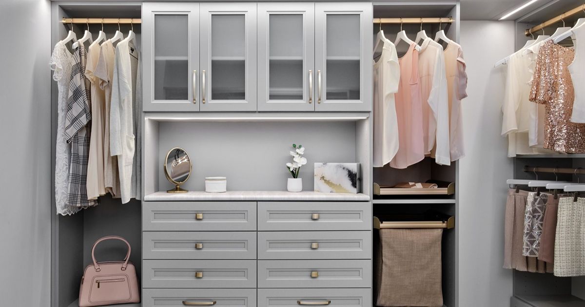 A modern, light gray walk-in closet with hanging clothes, glass-front cabinets, open shelving, and a set of drawers.