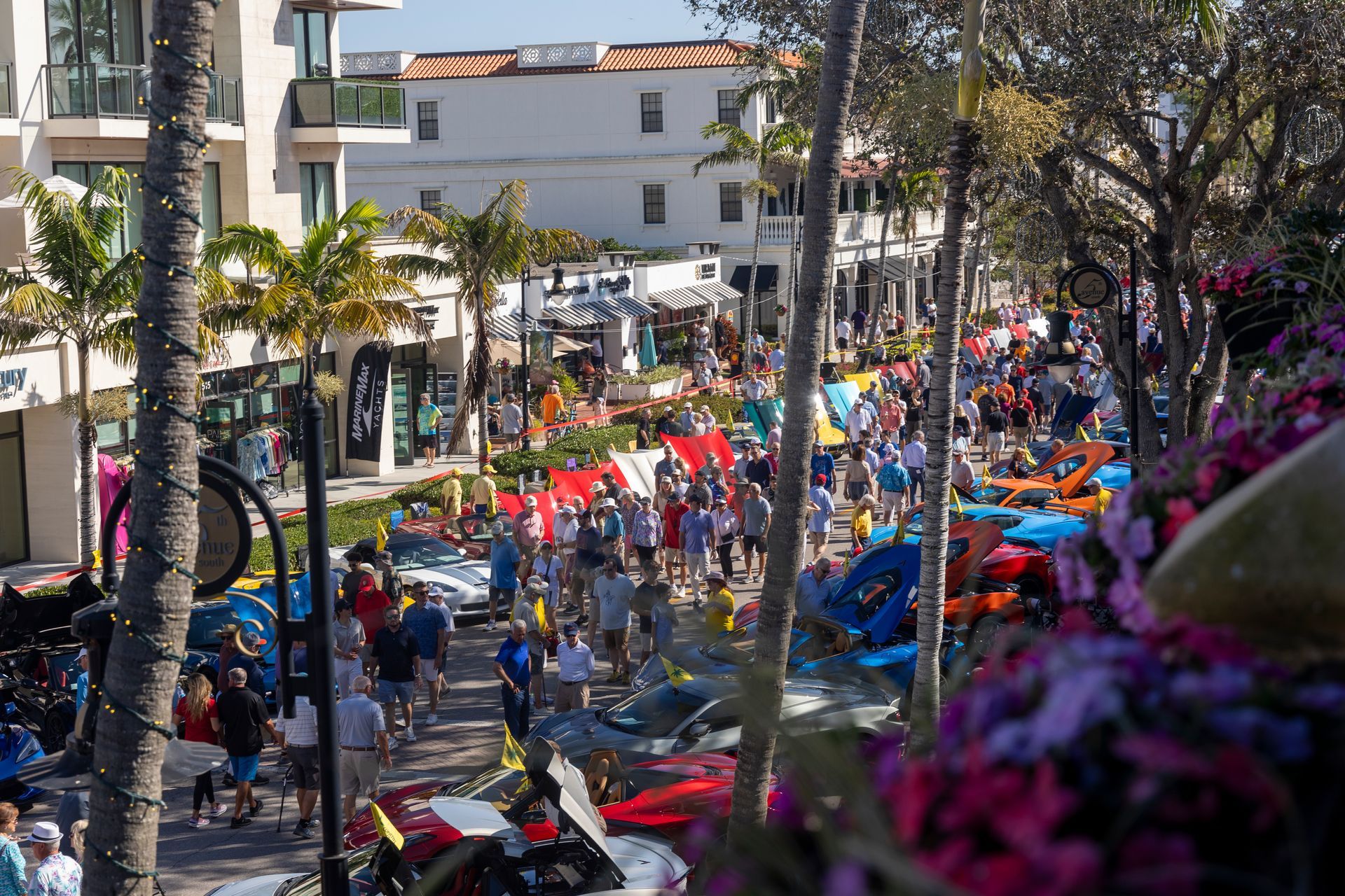 Crowd of people walking and milling around a street lined with shops and palm trees.