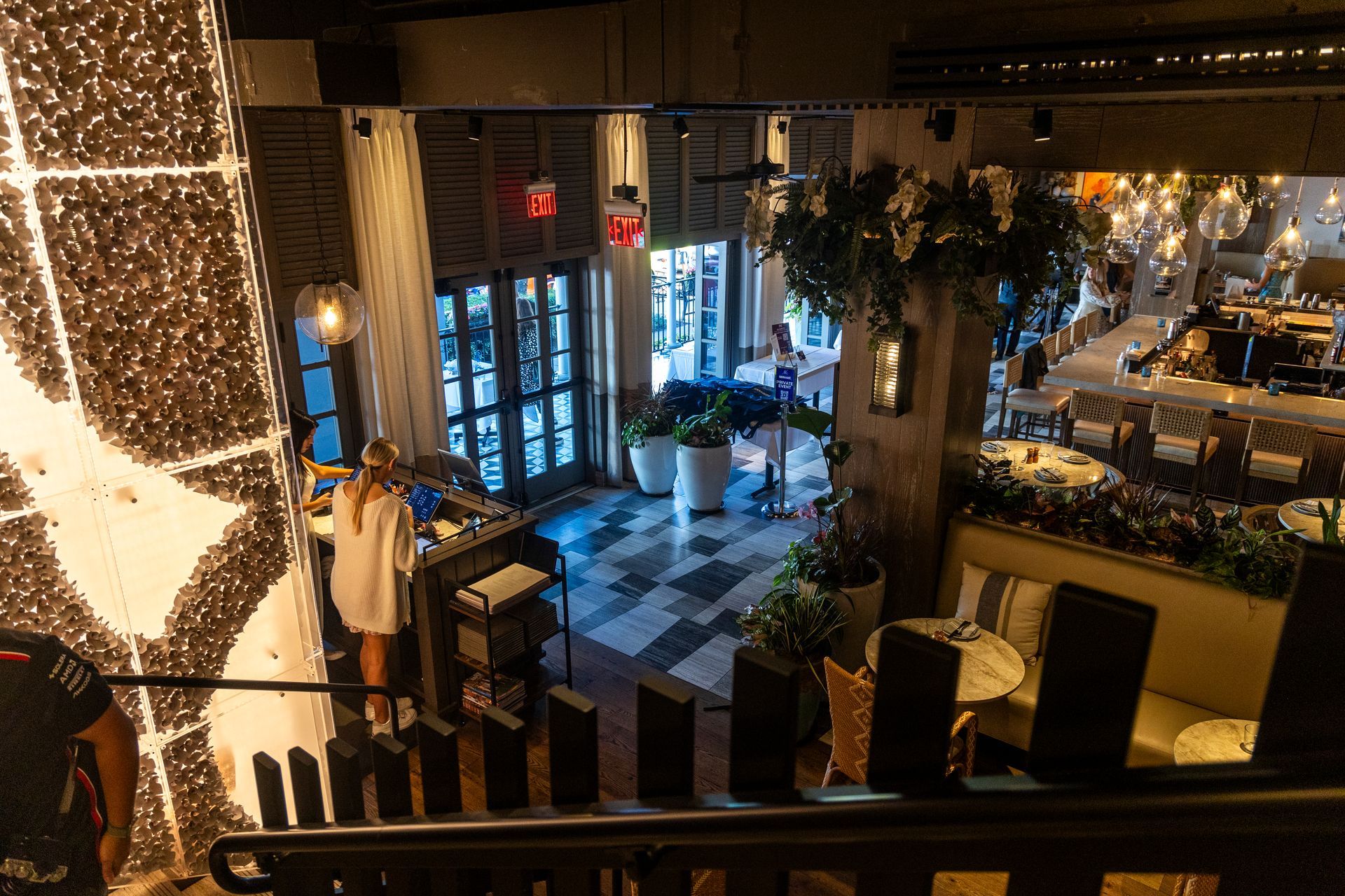 Restaurant interior with checkered floor, entrance, and staircase, seen from above. A person stands near the entrance.