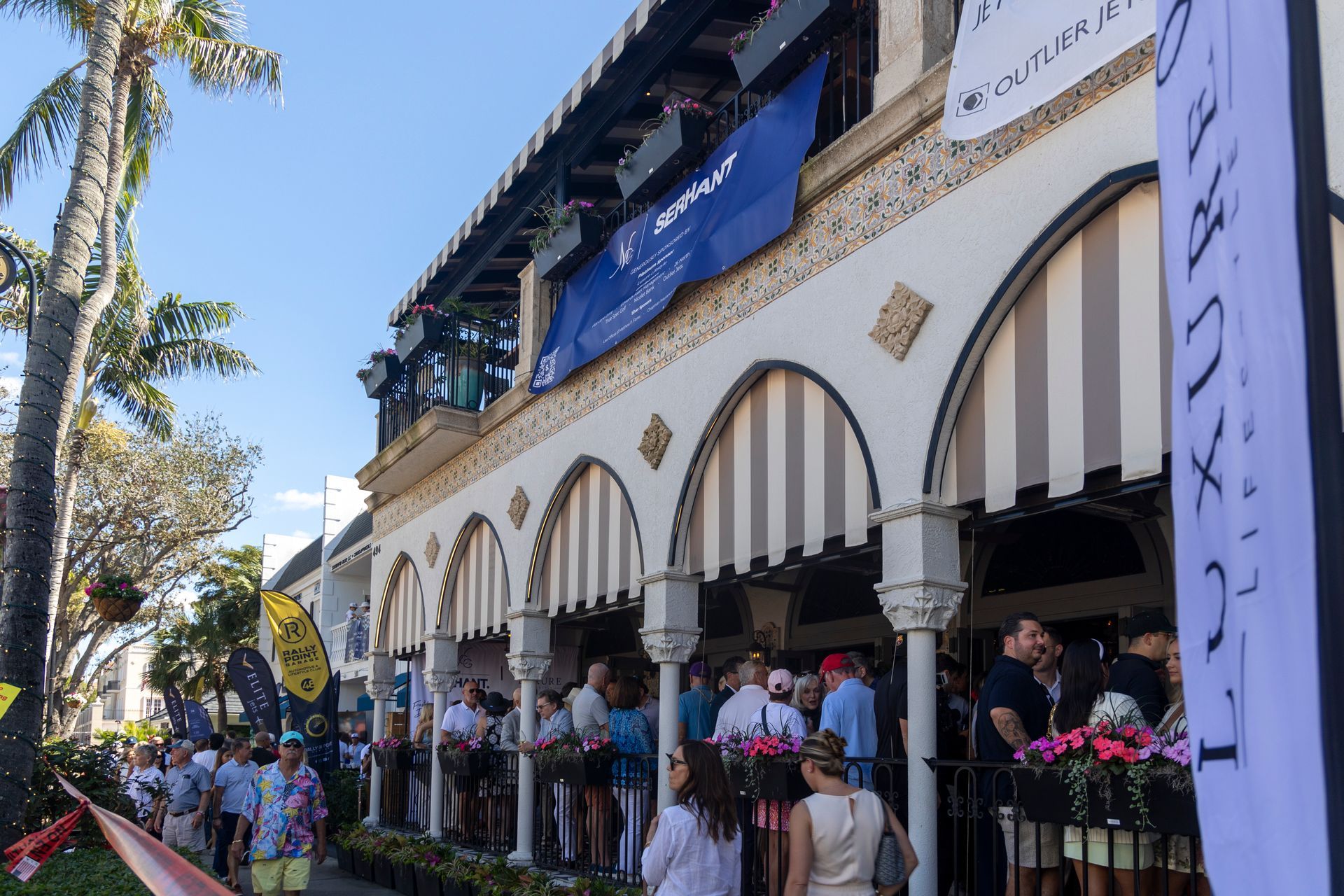 Exterior of a building with arches, awnings, and a crowd of people gathered outside on a sunny day.