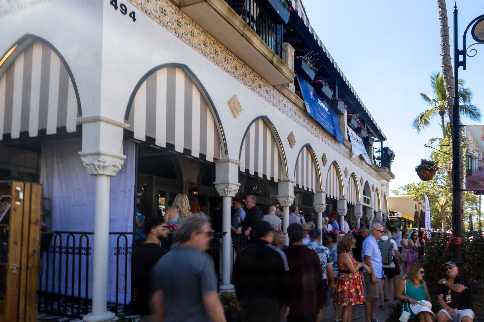 Building with arched windows and striped awnings, lined with people on a sunny street.