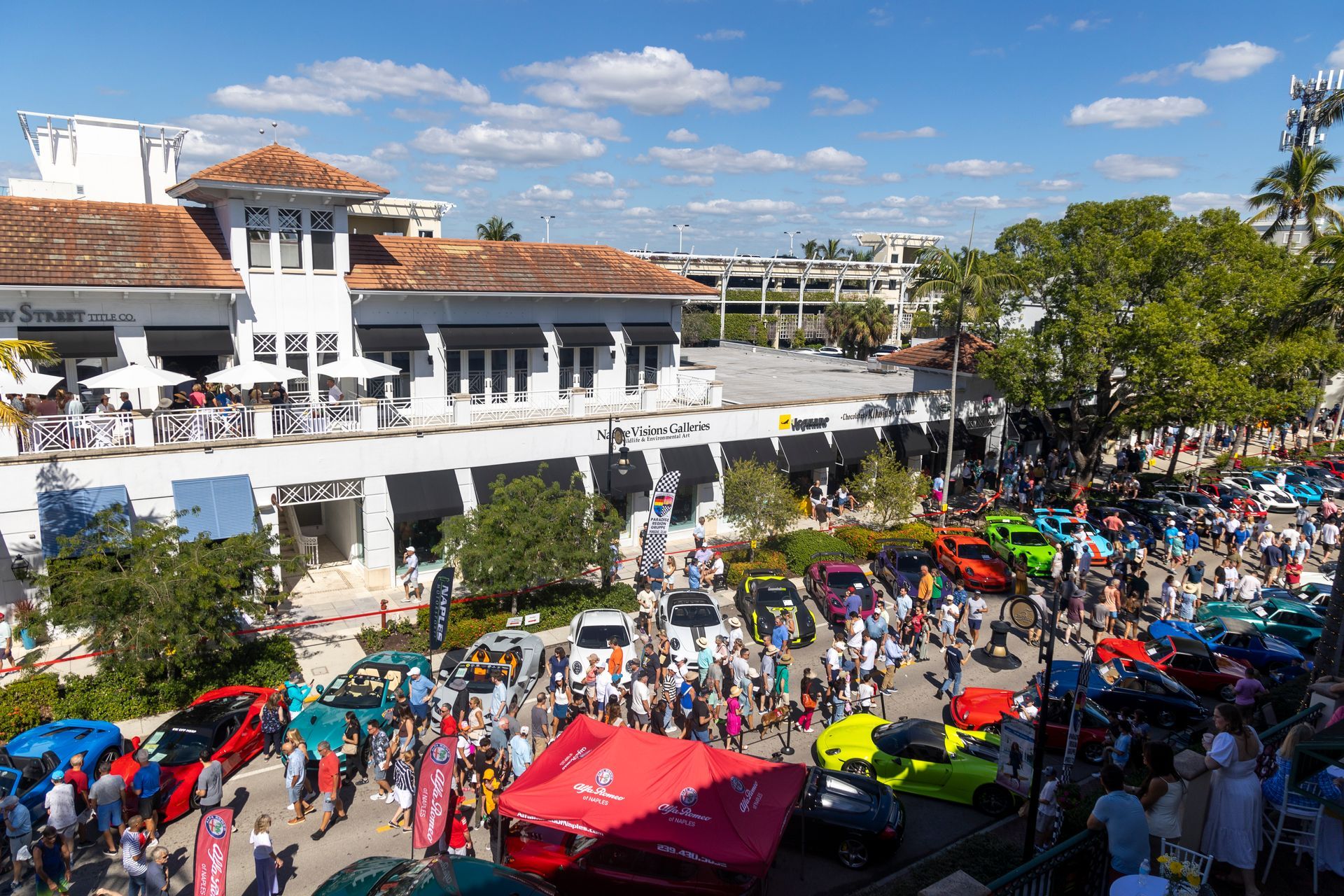 Crowd at car show with colorful vehicles and white buildings. Bright sunny day.