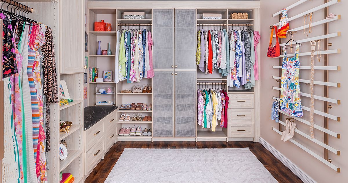 A organized walk-in closet with light wood shelving, hanging clothes, drawers, a central rug, and a wall accessory rack.