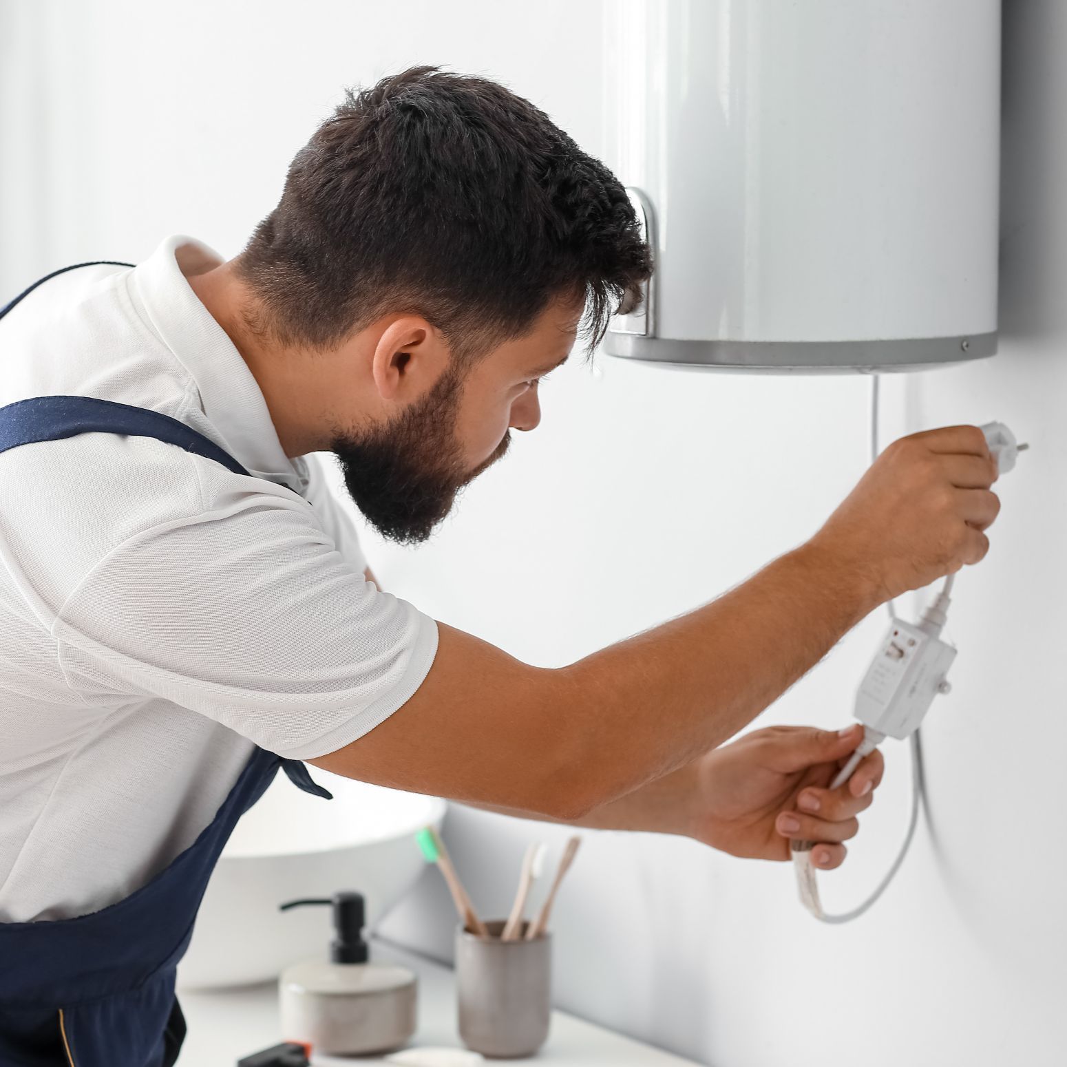 A person in work overalls plugging an electric water heater cord into a wall outlet in a bathroom.