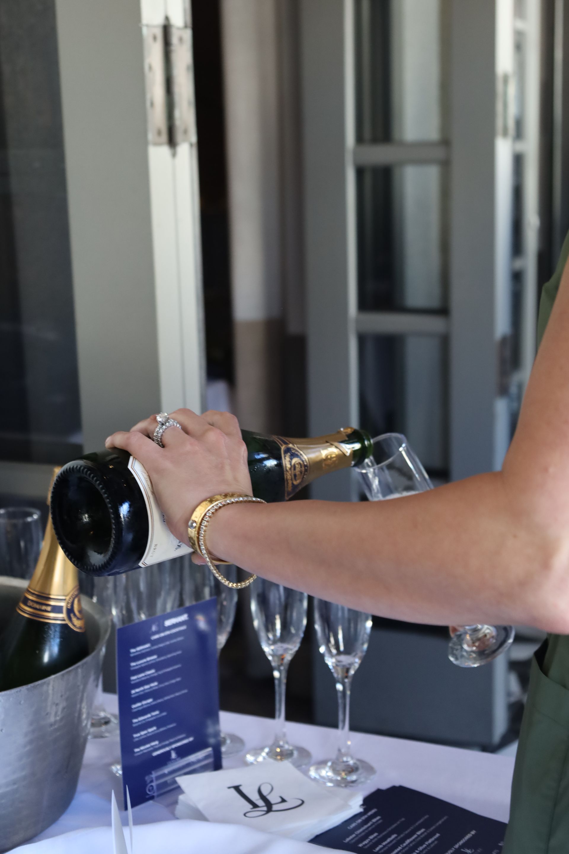 Woman pouring champagne into glasses at an outdoor event, near a bucket and a menu.