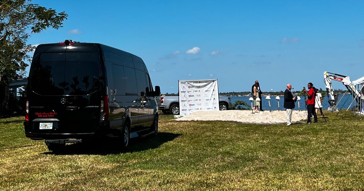 Black van parked on grass, people near a sandy area with a structure near a body of water.