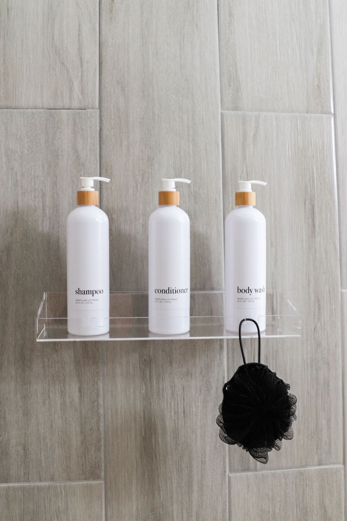 White shampoo bottles on a clear shelf in a shower, with a black loofah.