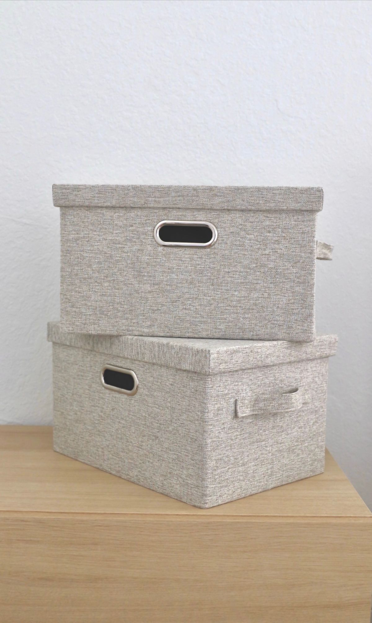 Two beige fabric storage boxes, one on top of the other, with handles, on a wooden surface against a white wall.
