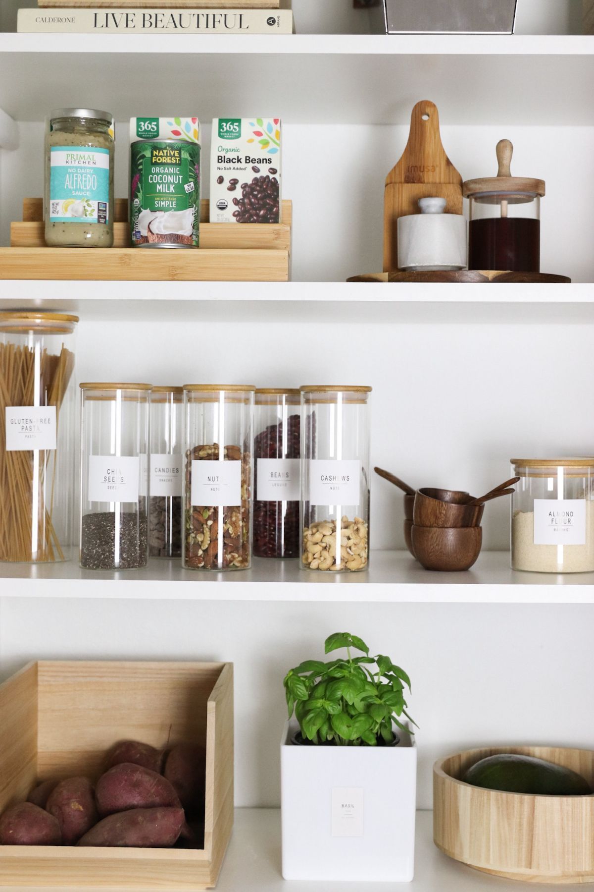 Organized pantry with various food items in jars, boxes, and a plant on white shelves.
