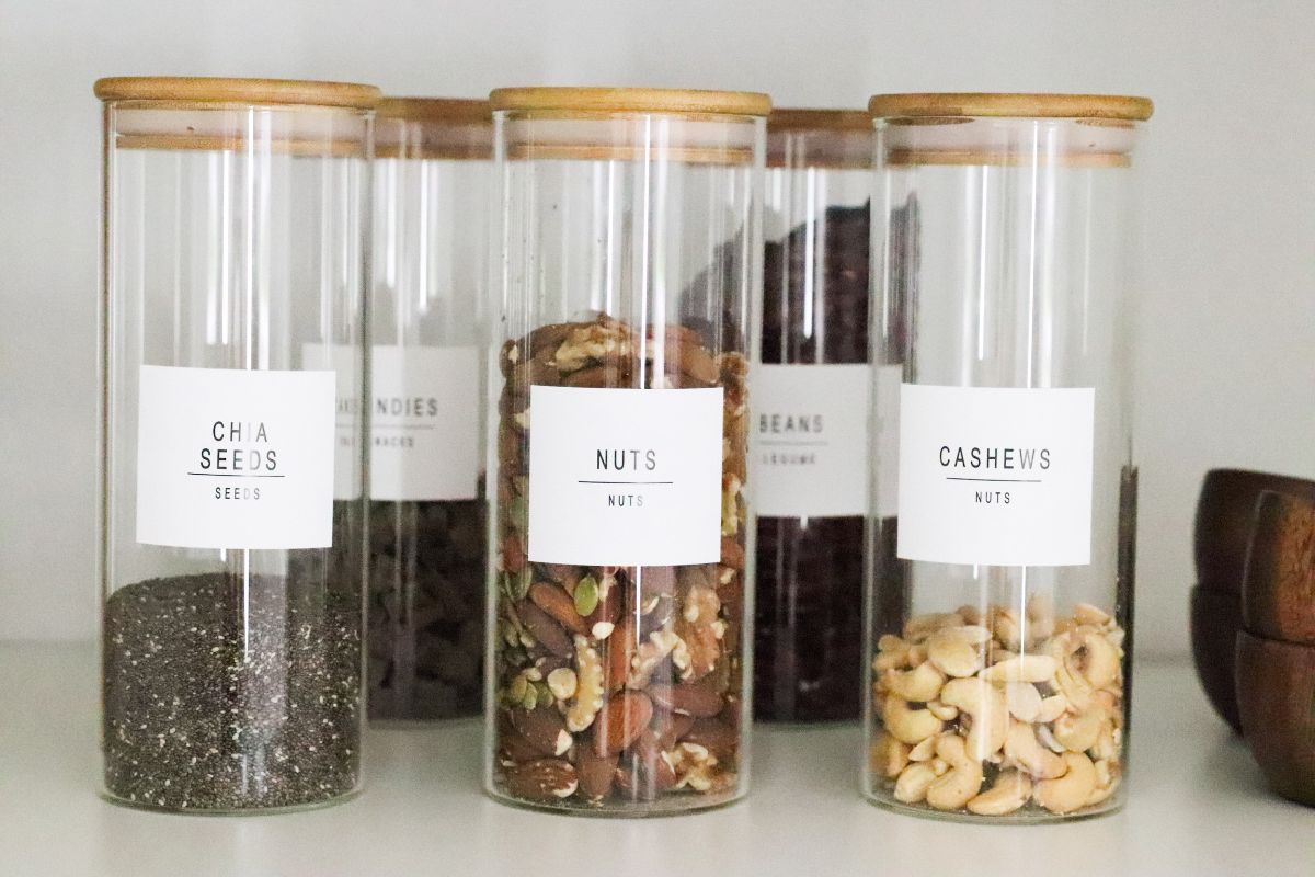 Clear glass jars with bamboo lids, each labeled and filled with various nuts and seeds, neatly arranged on a shelf.