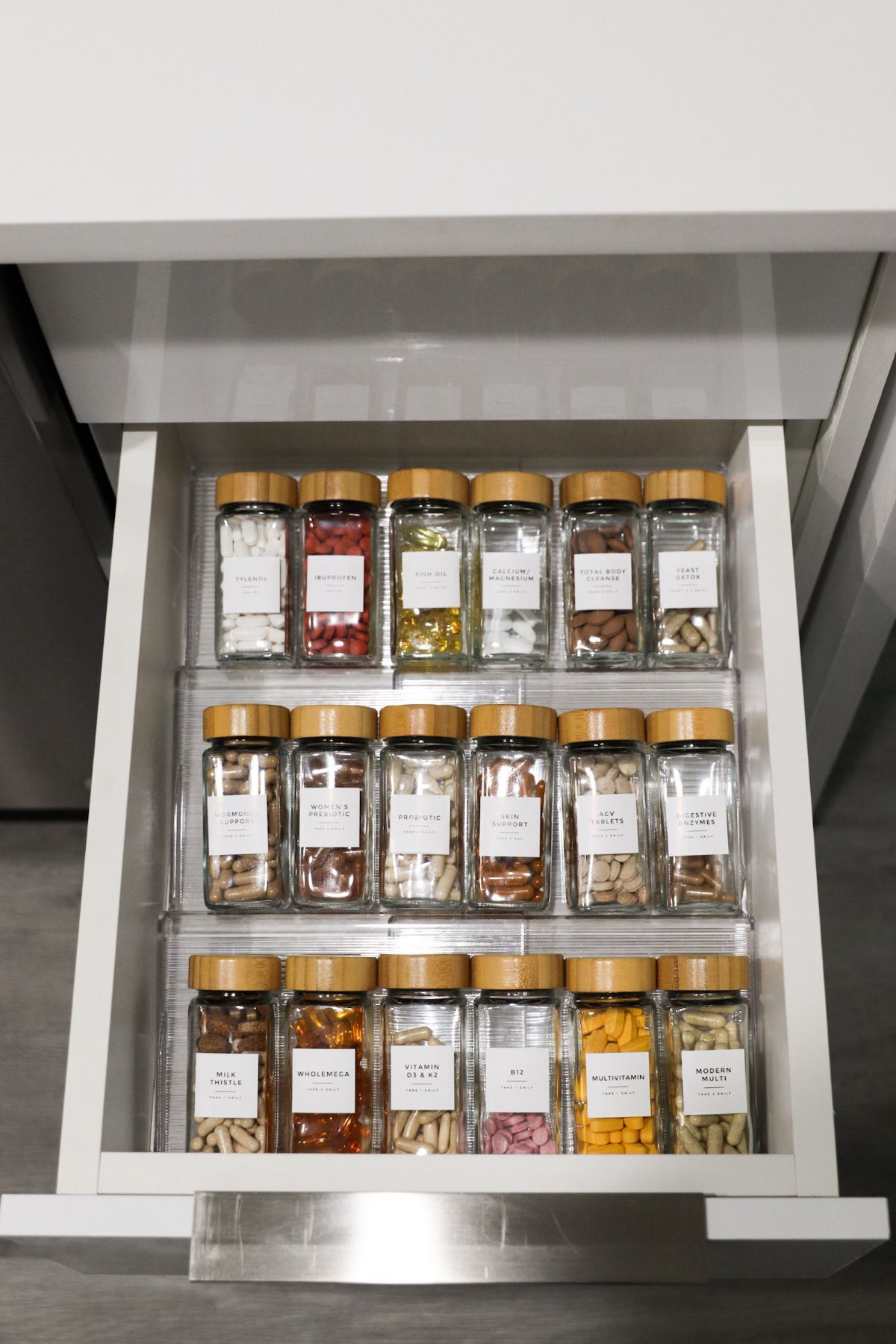 Spice jars neatly organized in a drawer. Clear glass jars with wooden lids and labels, arranged in three rows.