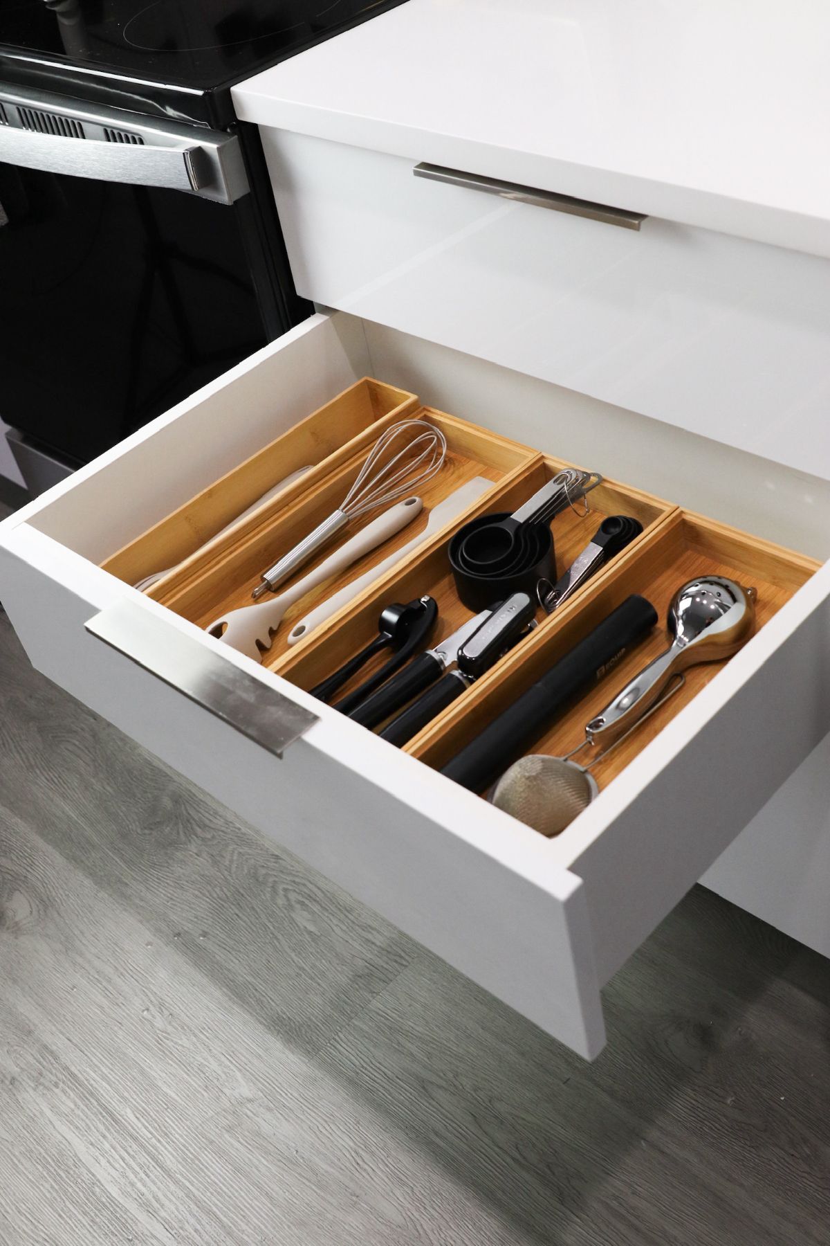 Open kitchen drawer with bamboo utensil organizer; white cabinet; stainless steel handle; various cooking utensils.