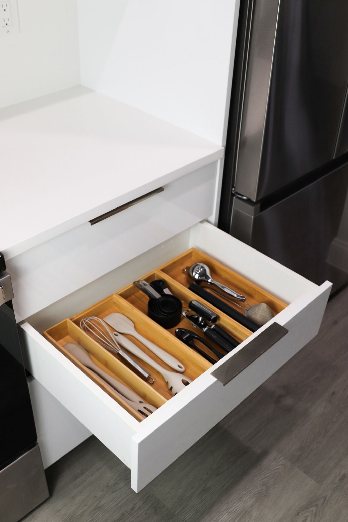 Open kitchen drawer with bamboo utensil organizer. White drawer, stainless steel appliances, light wood organizer.