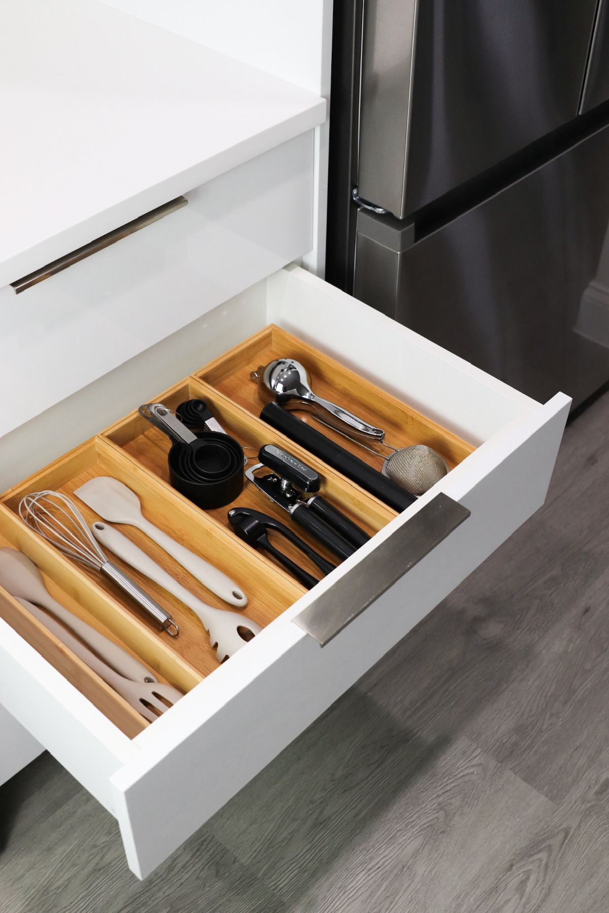 White kitchen drawer with bamboo utensil organizer, open. Silver handles, stainless steel fridge.