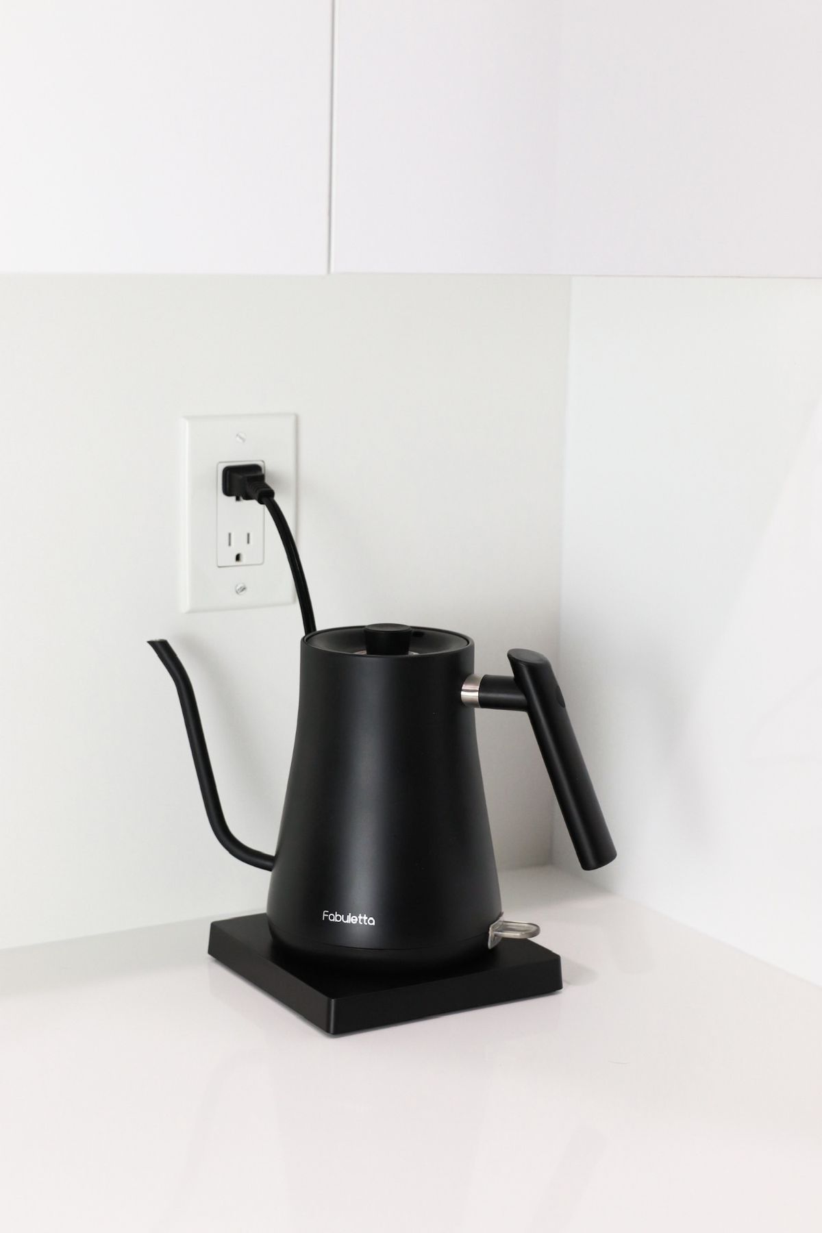 Black electric kettle on a stand, plugged into a wall outlet on a white counter.