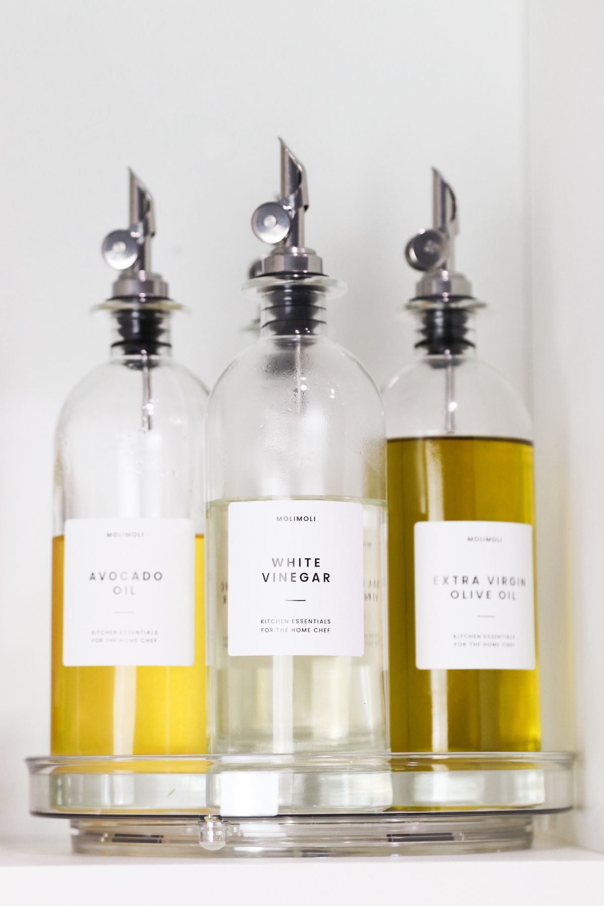Three glass bottles with silver spouts holding avocado oil, white vinegar, and extra virgin olive oil on a shelf.